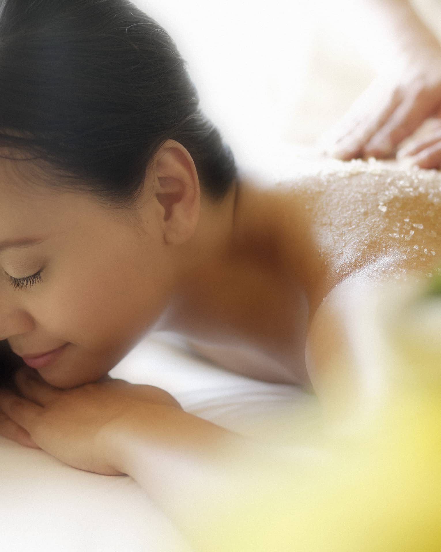 Woman lying down with eyes closed as hands massage salt scrub into shoulders