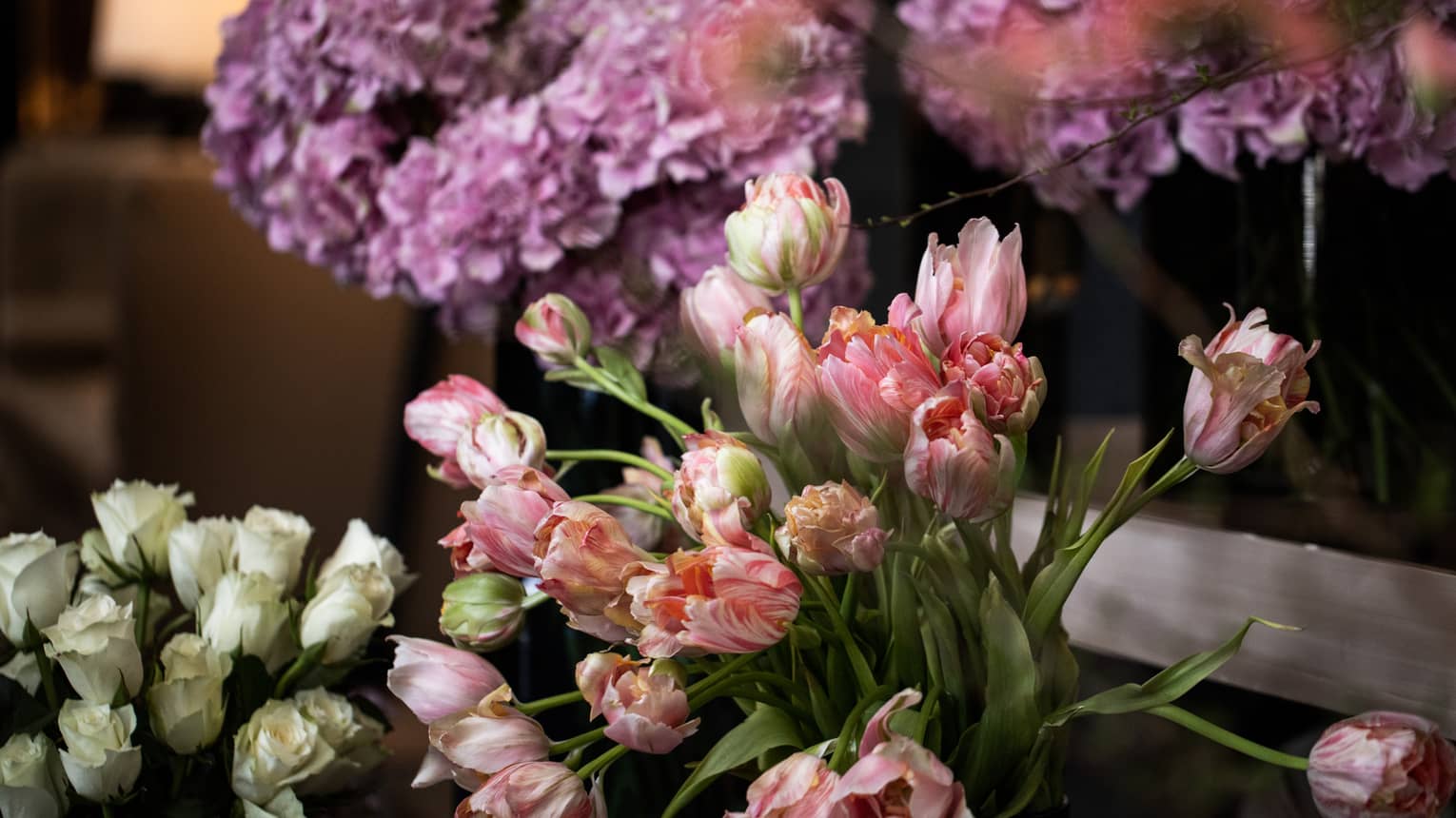 Three separate bouquets of light-purple hydrangeas, pink tulips and white roses sit gathered on a tiered table