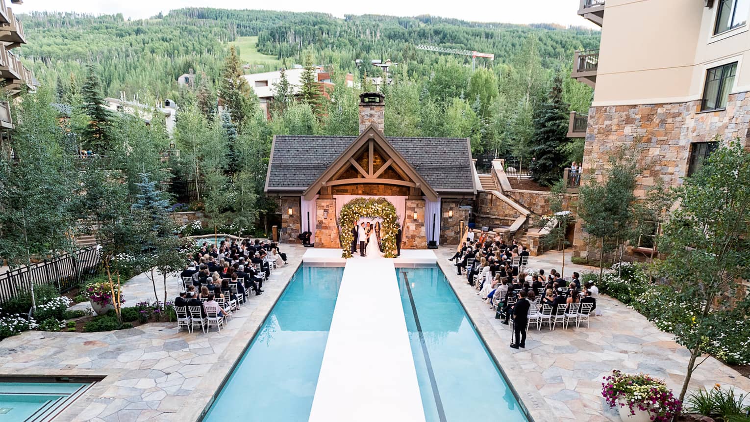 Aerial view of rows of wedding guests face bride and groom on pool terrace, mountains 