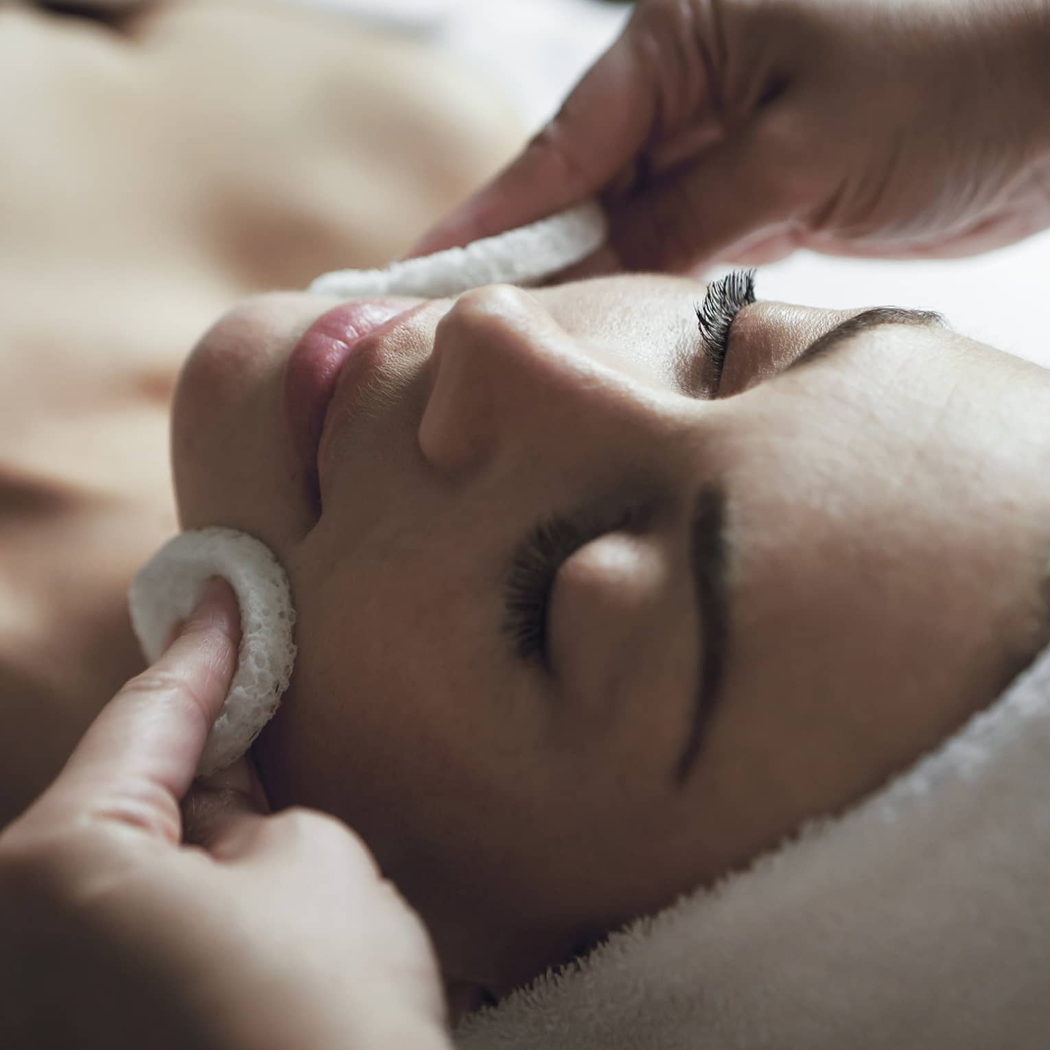 A woman with relaxes as a spa professional uses two sponges to exfoliate her face