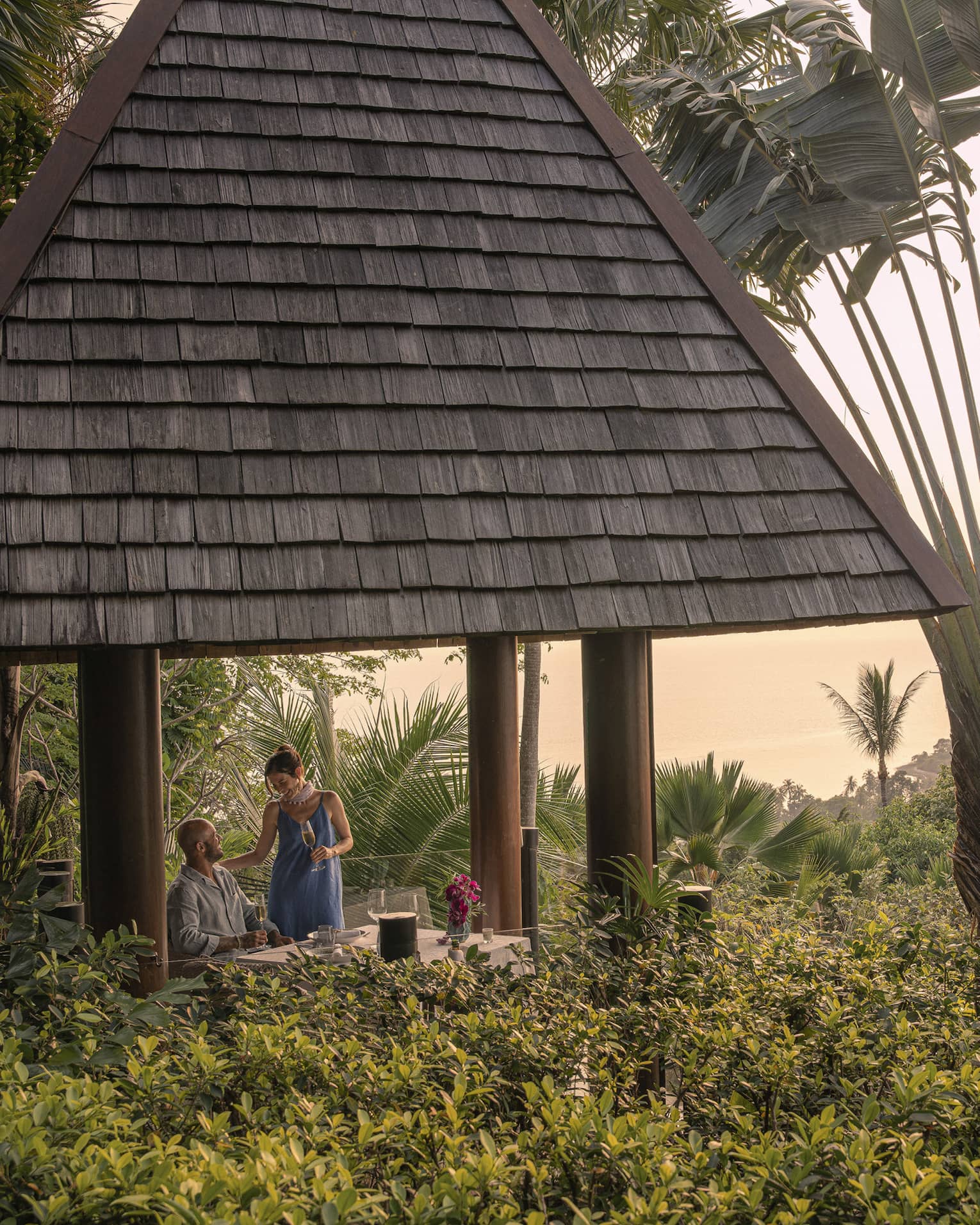 A couple with champagne in a tall hilltop pavilion with a dark, straw roof, set amidst lush forest and a giant fan palm tree.