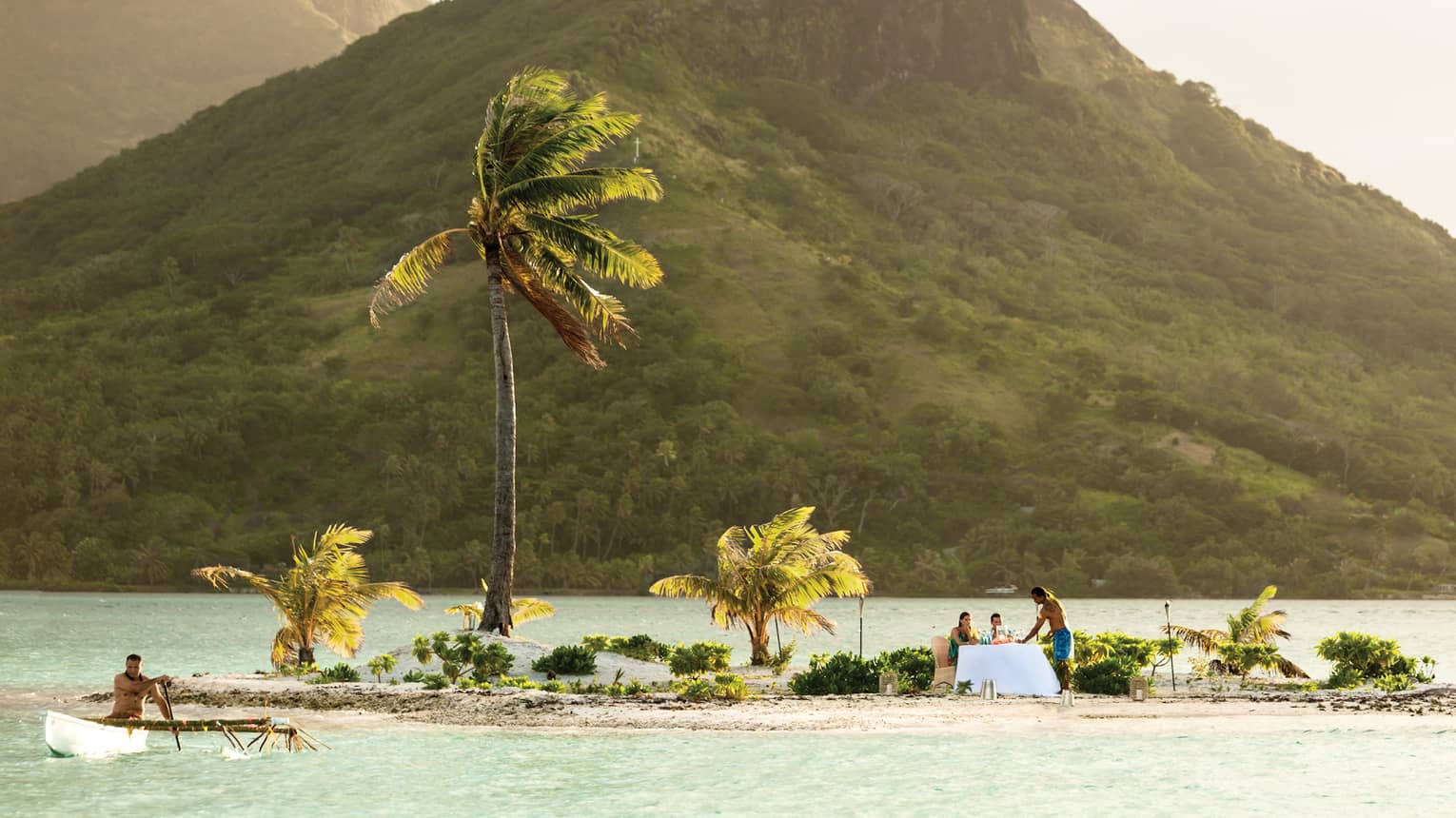 Couple dines on small private island with windswept palm trees in lagoon under mountain