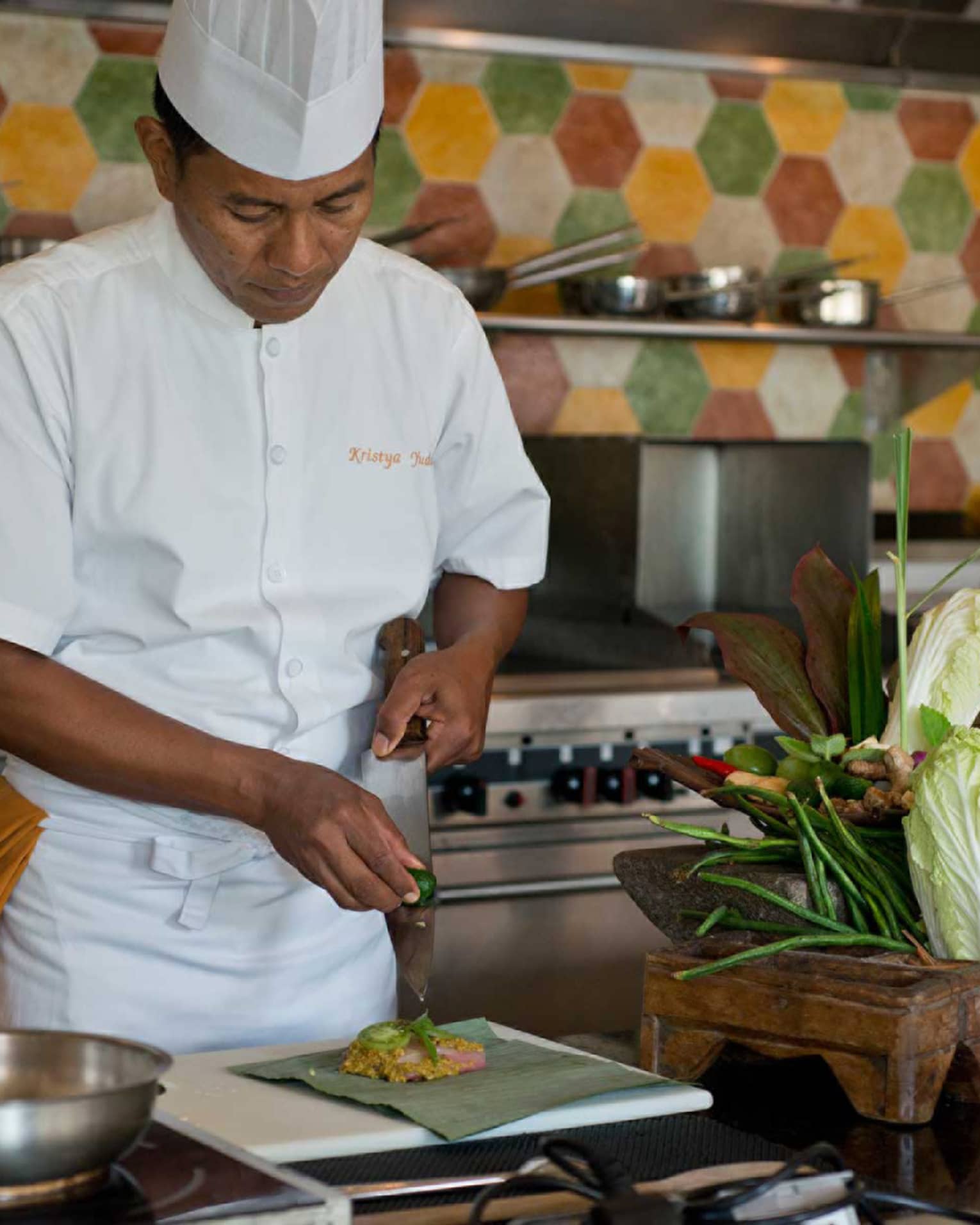 Chef in white uniform squeezes lime on large kitchen knife over dish, next to heads of fresh lettuce and whole peppers