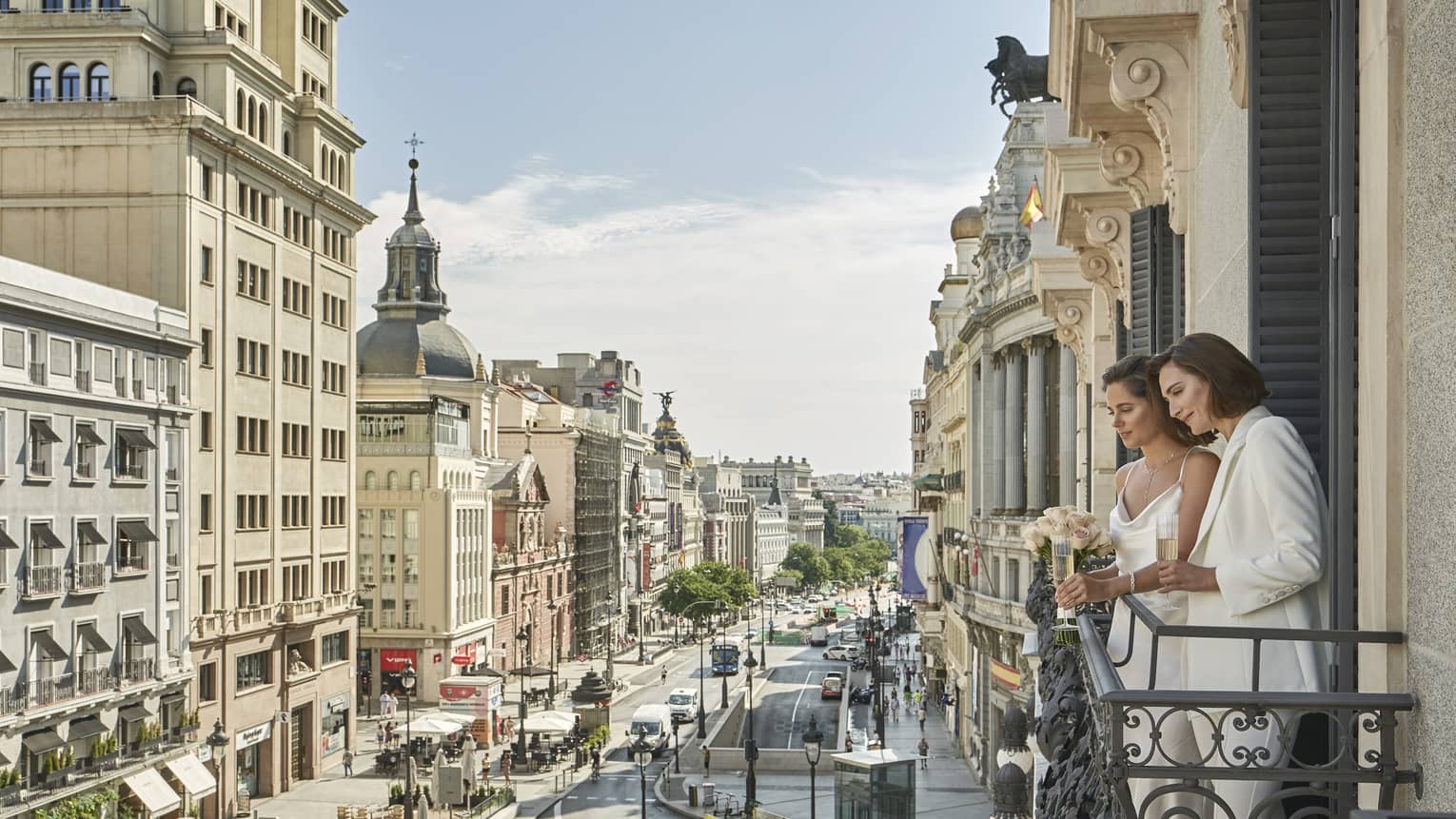 Two women in white on a balcony look out over the bustling street of Madrid surrounded by historical buildings.