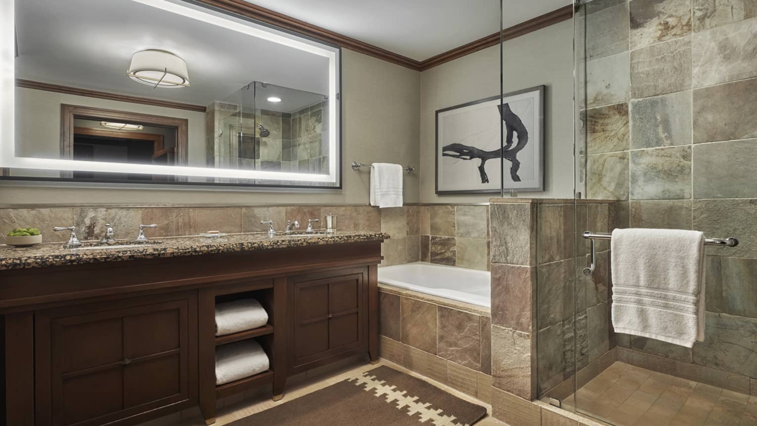 A tiled bathroom, with a double vanity, large lighted mirror and a shower and tub separated by a glass partition