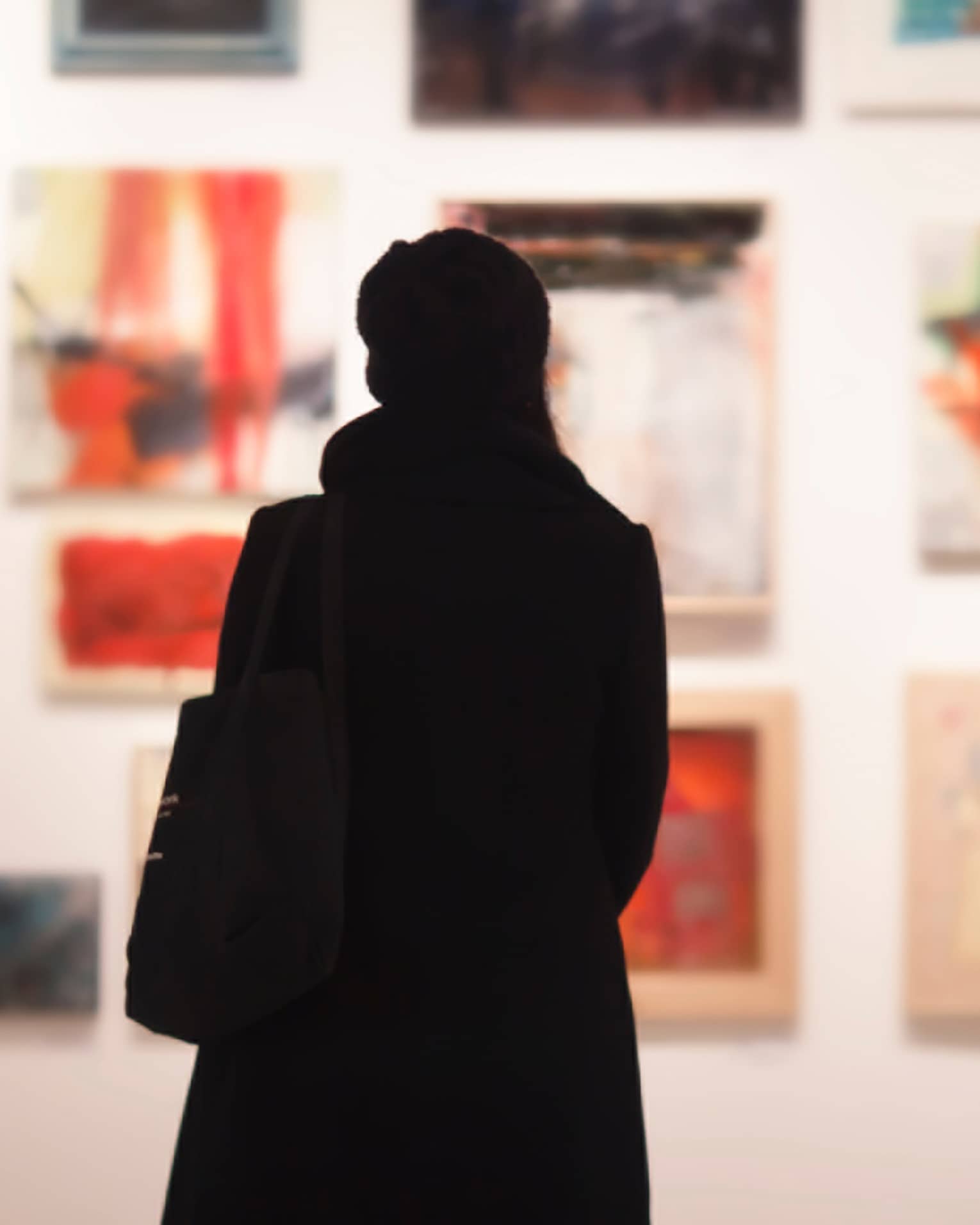 Silhouette of back of woman with hat bag looking at gallery wall with colourful art