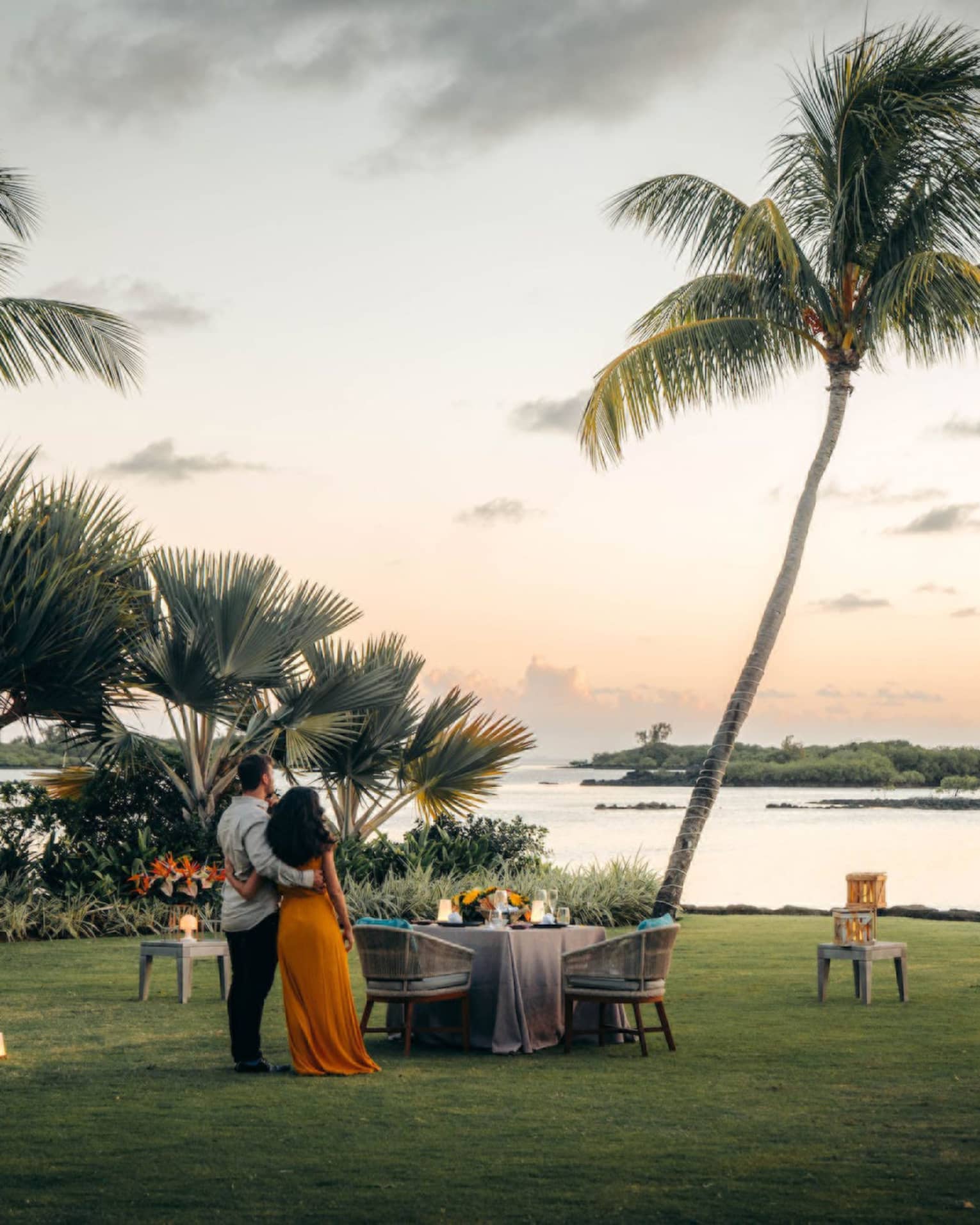 A couple stand on the shore, arms around each other, gazing toward the ocean; set table with two chairs beside swaying palms.