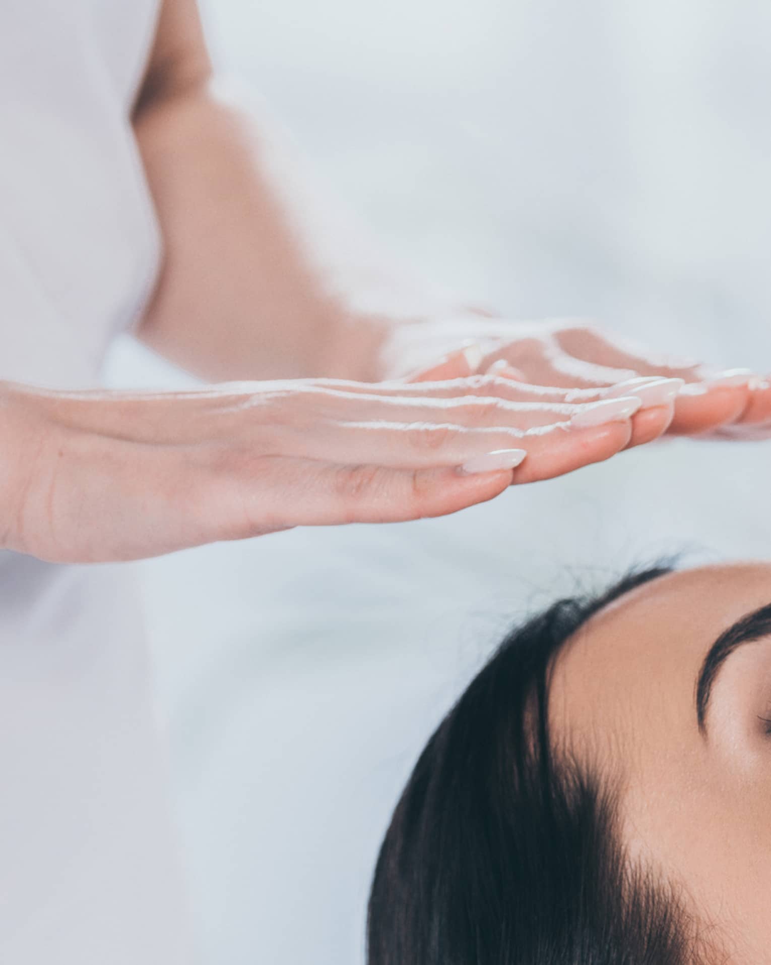 Woman with eyes closed lays on rolled up towel on spa table under therapists' hovering hands