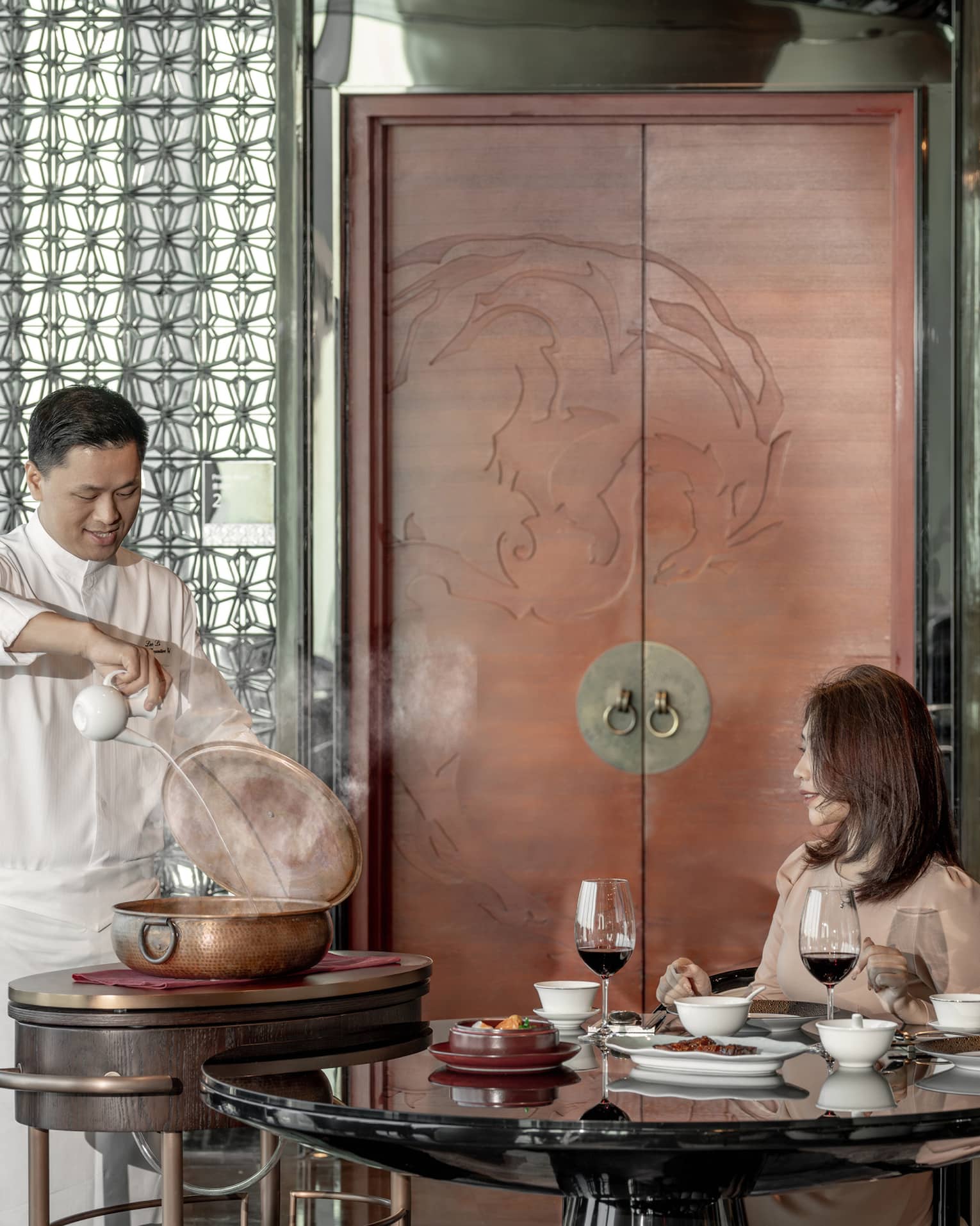 Two female diners seated at table watch as chef pours liquid into large copper pot 