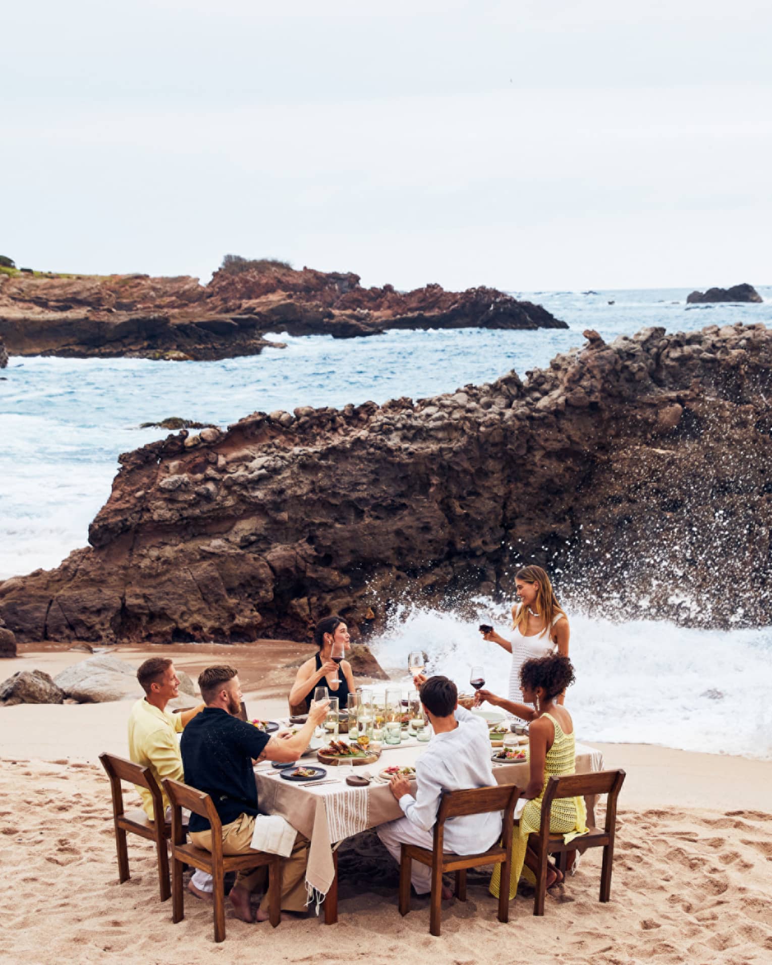 A group eating at a table on a beach with waves crashing nearby.