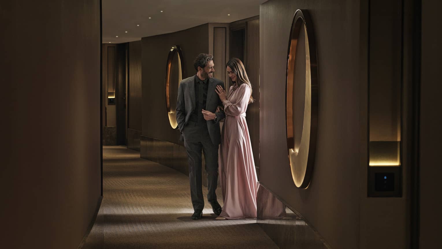 Smiling couple in formal wear stands in a dimly lit hall with oversized oval mirrors