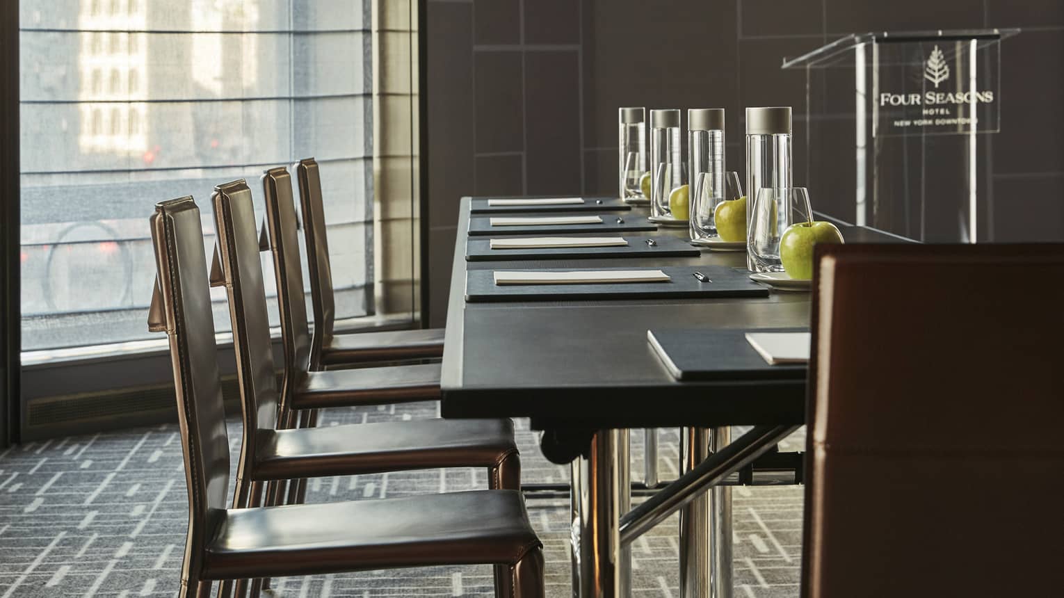 Dark table with chairs, stationery, bottles of water, green apples in light-filled meeting room with podium