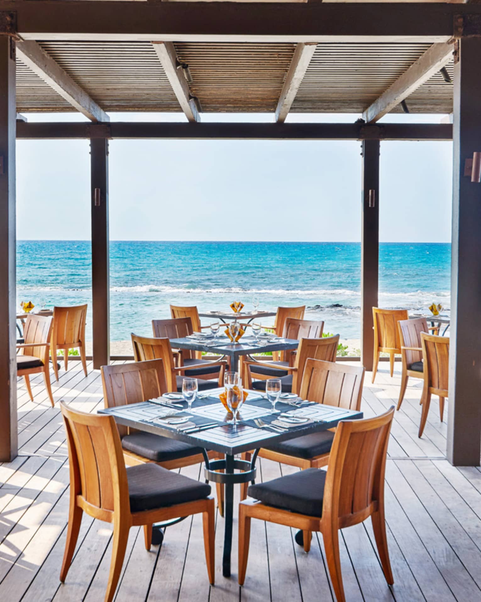  The Ulu Ocean Grill and Sushi Lounge is ready for guests with square tables set with menus and clear glassware, and overlooking the ocean from the covered wooden terrace. 