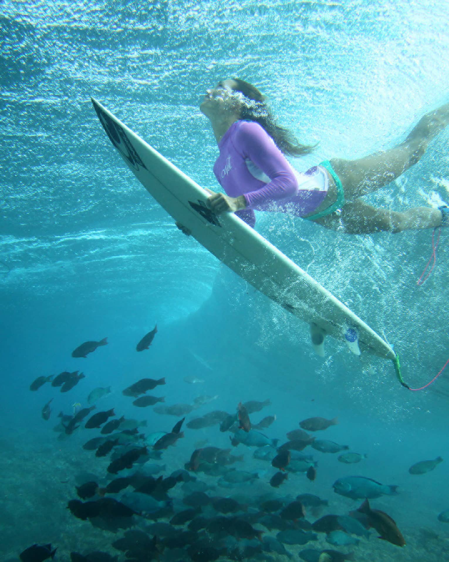 Céline Gehret, ascending from the water above a school of fish as she surfs in the Maldives