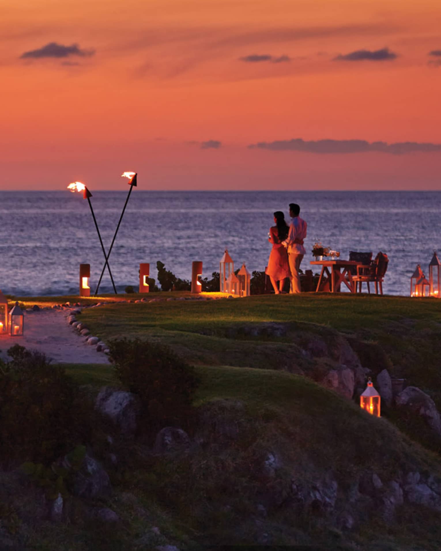 Couple stands on The Rock outpost at dusk, overlooking ocean with private dining table with lanterns, torches 