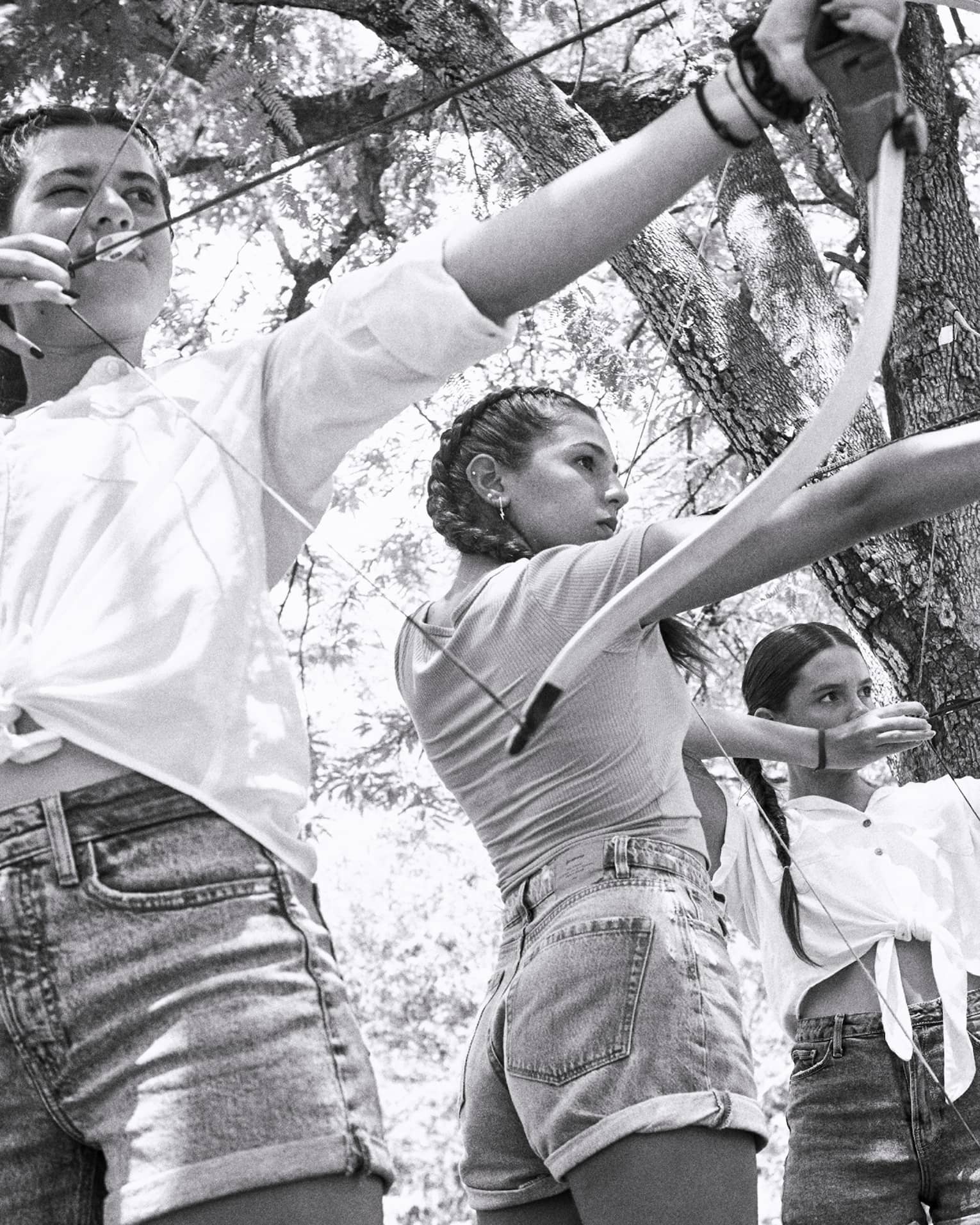 Black and white photo of three young girls aiming traditional archery bows in a forest