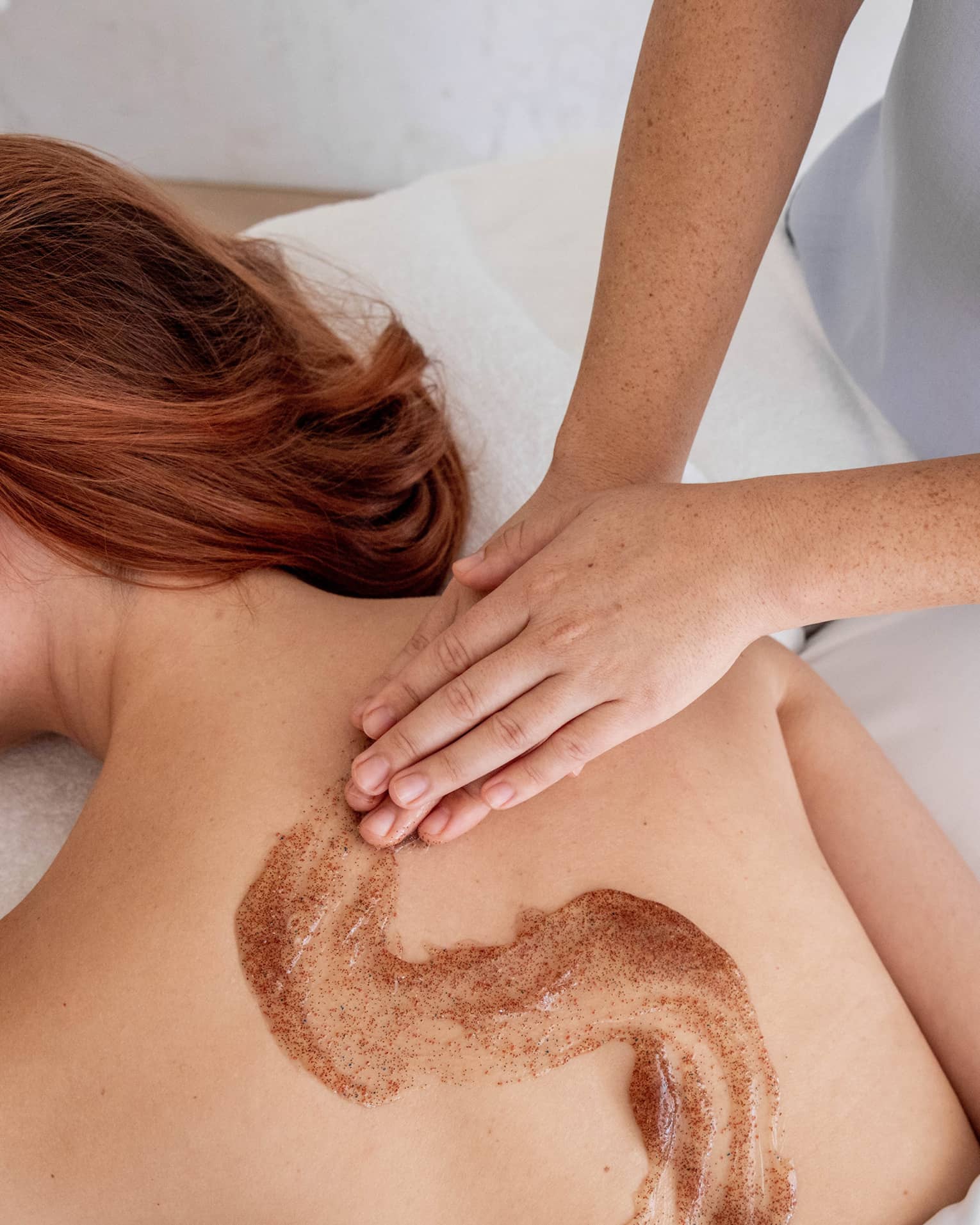 Spa staff massages scrub onto woman's bare back in treatment room