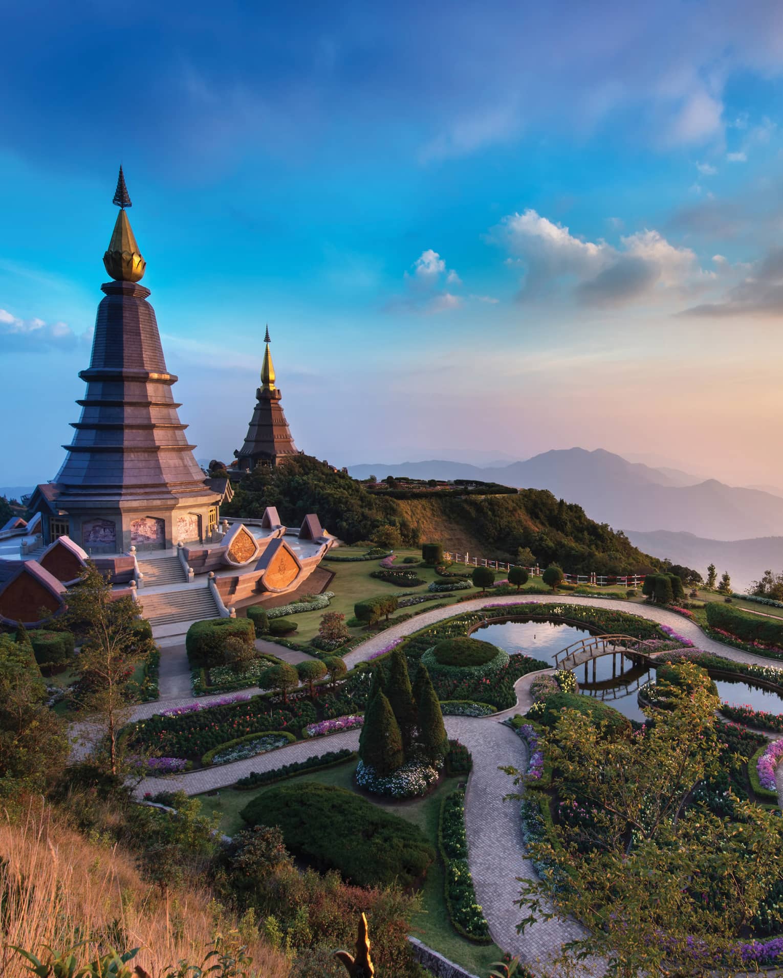 Two domed chedi monuments flanked by wending paths and manicured foliage atop a mountain, rose and gold sky and hills beyond.