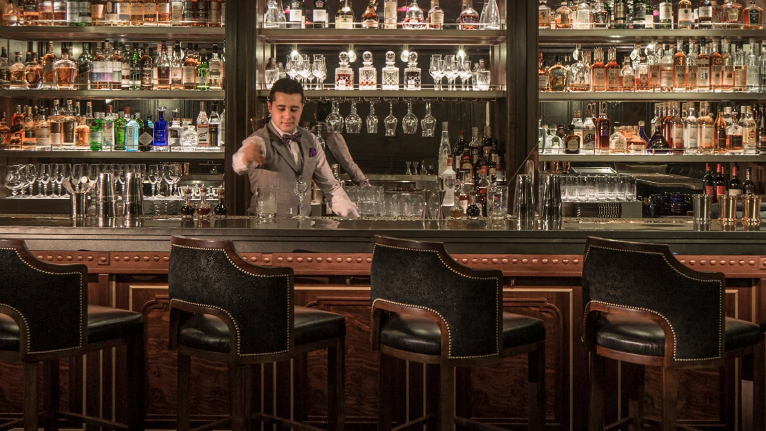 SIRR bartender in vest stands behind bar stirring cocktail, glasses and bottles lining wall behind him