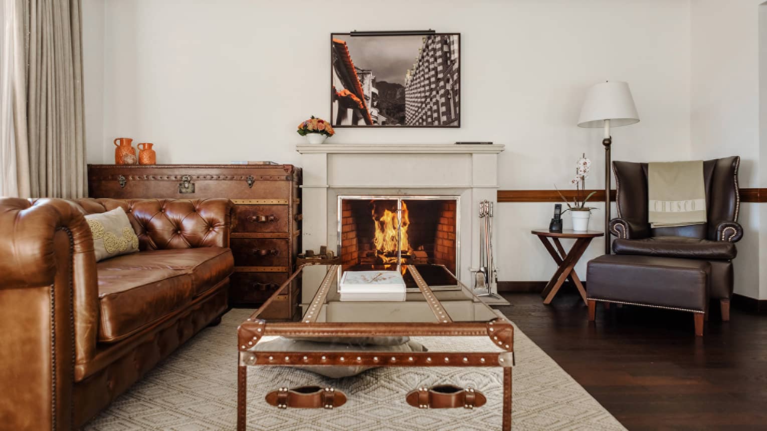 A brown leather chesterfield sofa, dark-brown leather arm chair and two trunk-style tables frame a central fireplace in the living room