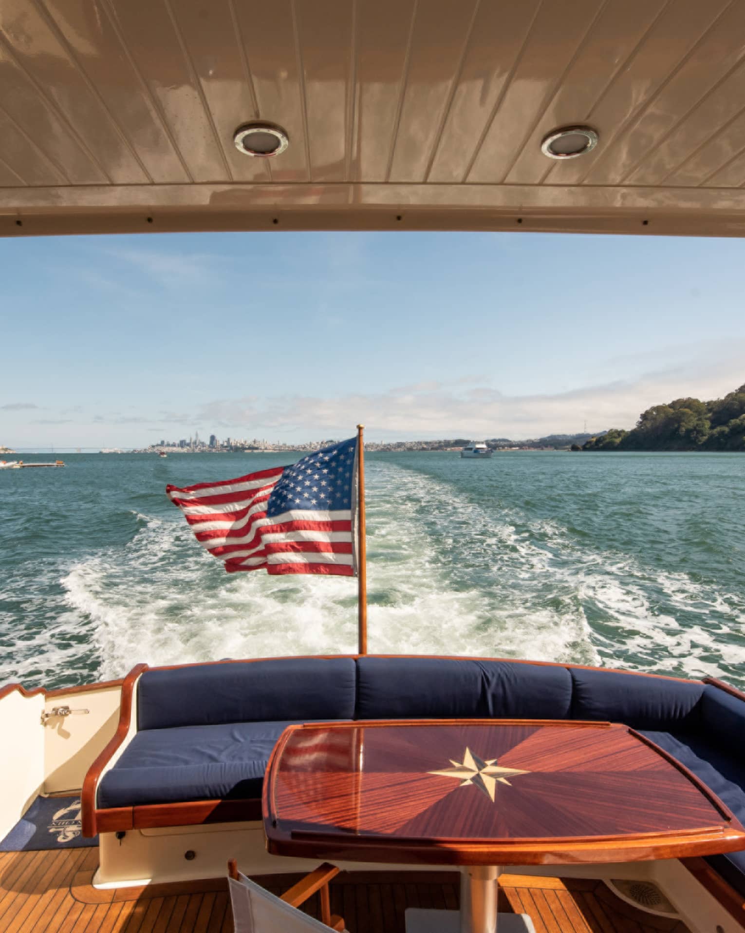 The back of a yacht, with a red table and an American flag, moving along the water.