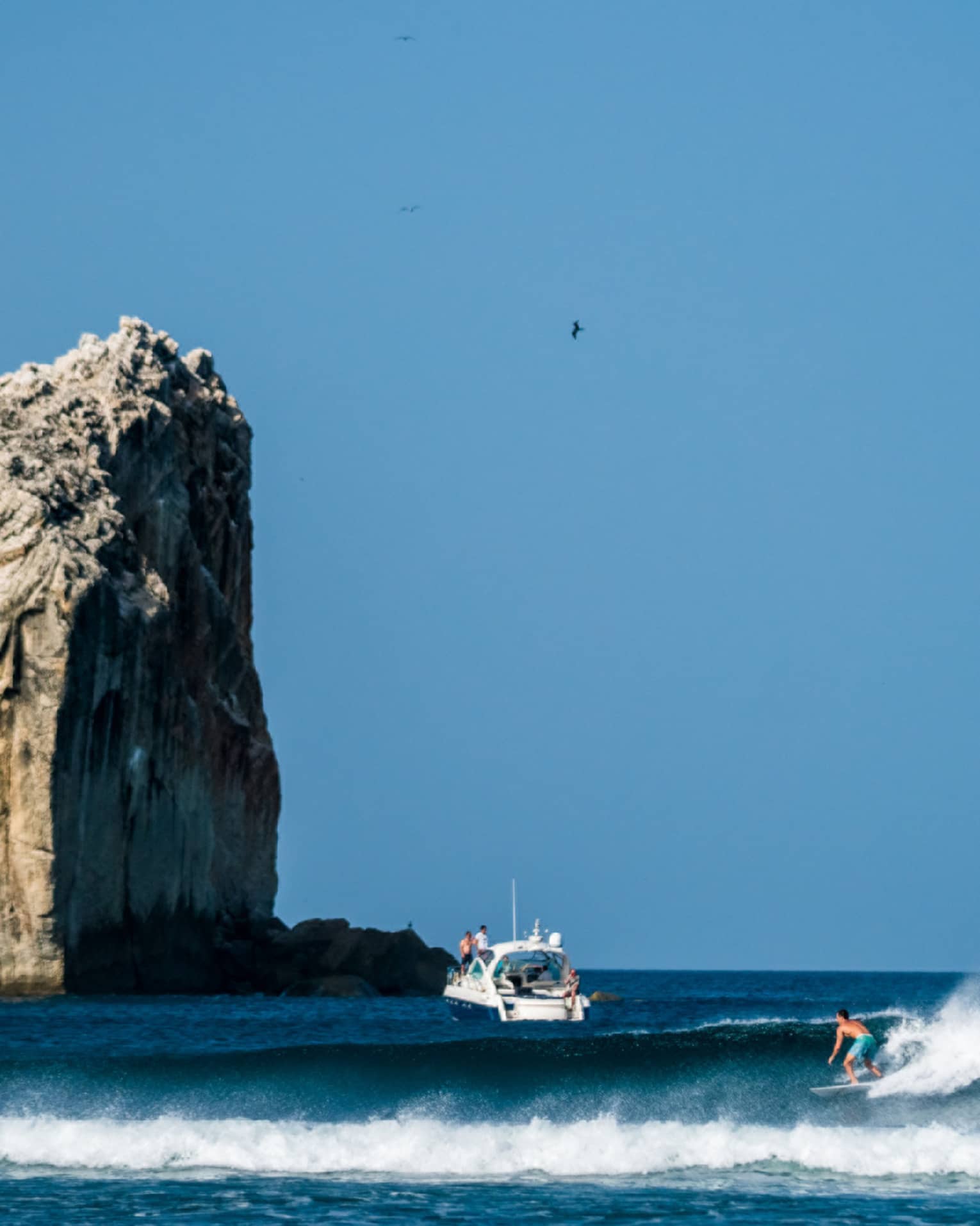 A surfer rides a wave in the foreground, with a boat nearby and a large rock formation towering above the water in the background