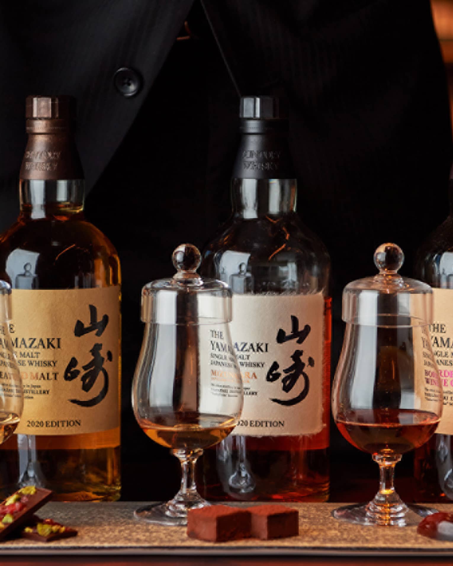 Hands frame a row of three bottles of single malt Japanese whisky behind three curved whisky tumblers with tasting caps.