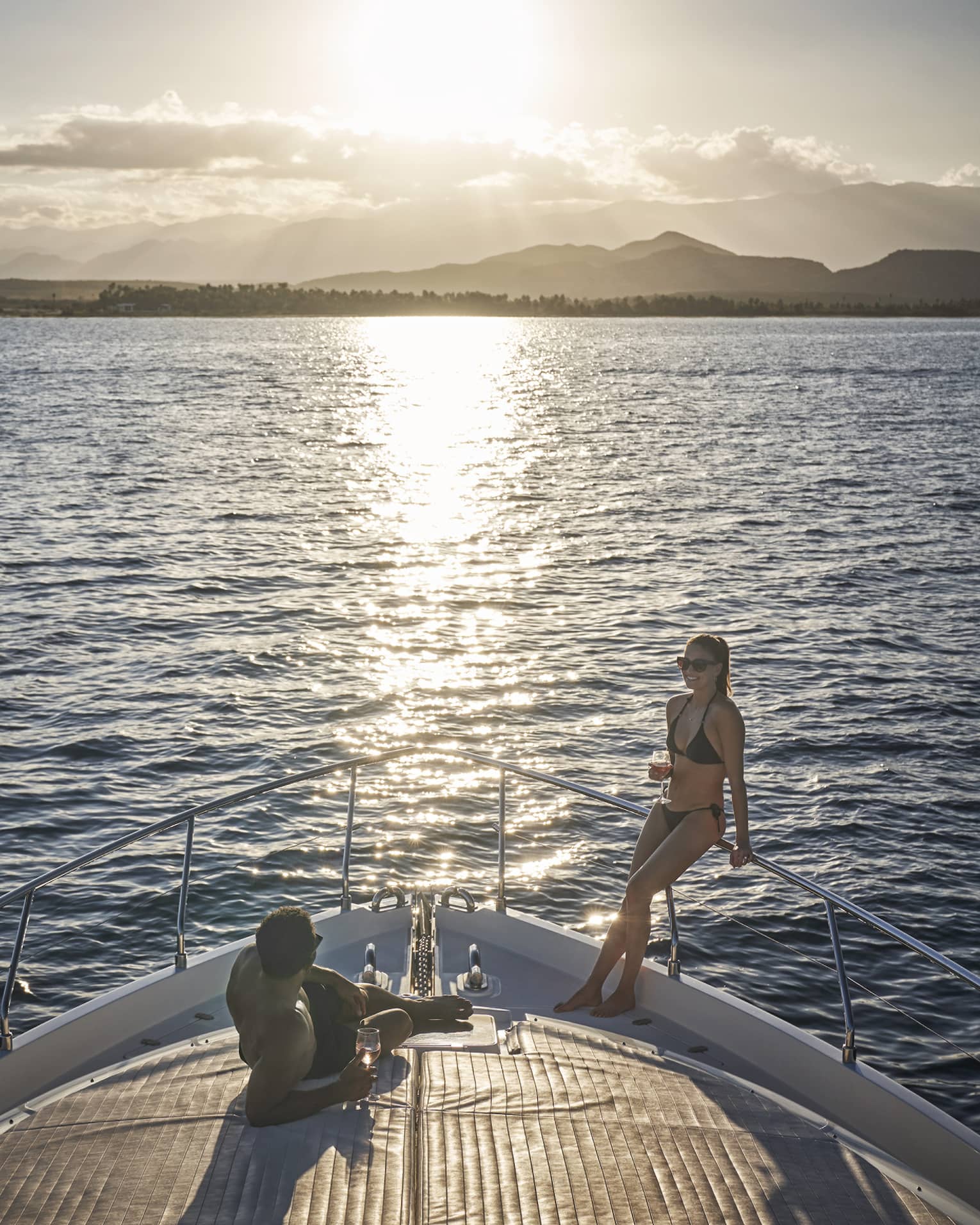 At the bow of a luxurious boat, a couple in swimwear lounges while enjoying the sun's glowing reflection and a glass of wine.