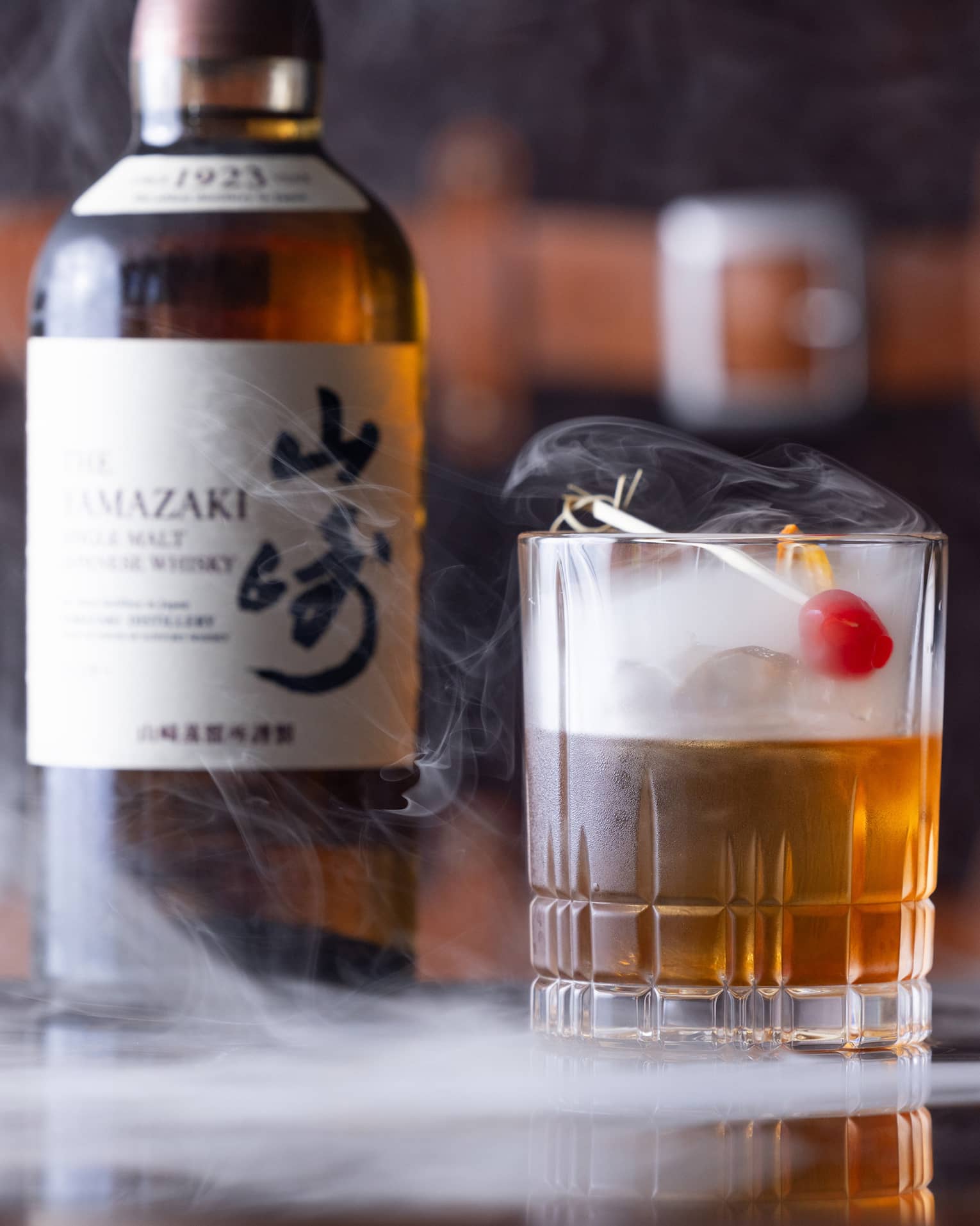Bottle of Japanese whisky beside half-full cut-crystal glass with red cherry garnish, dry ice mist swirling from the glass.