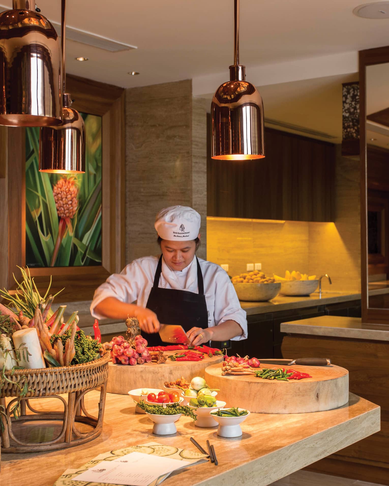 A chef chops vegetables on a large butcher block amid a vibrant array of fresh produce in a luxurious, warm-hued kitchen.