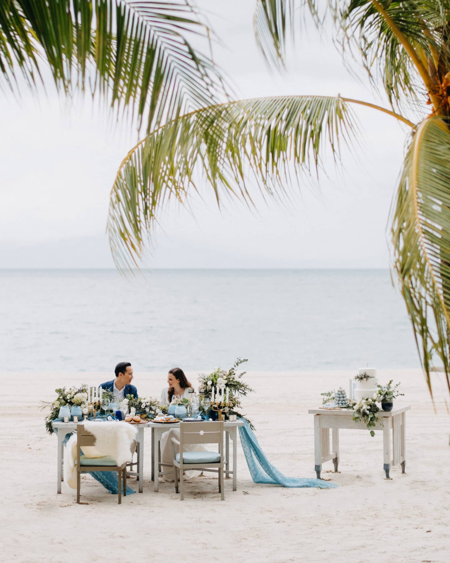 Smiling couple at private dining on white sand beach past palm leaves