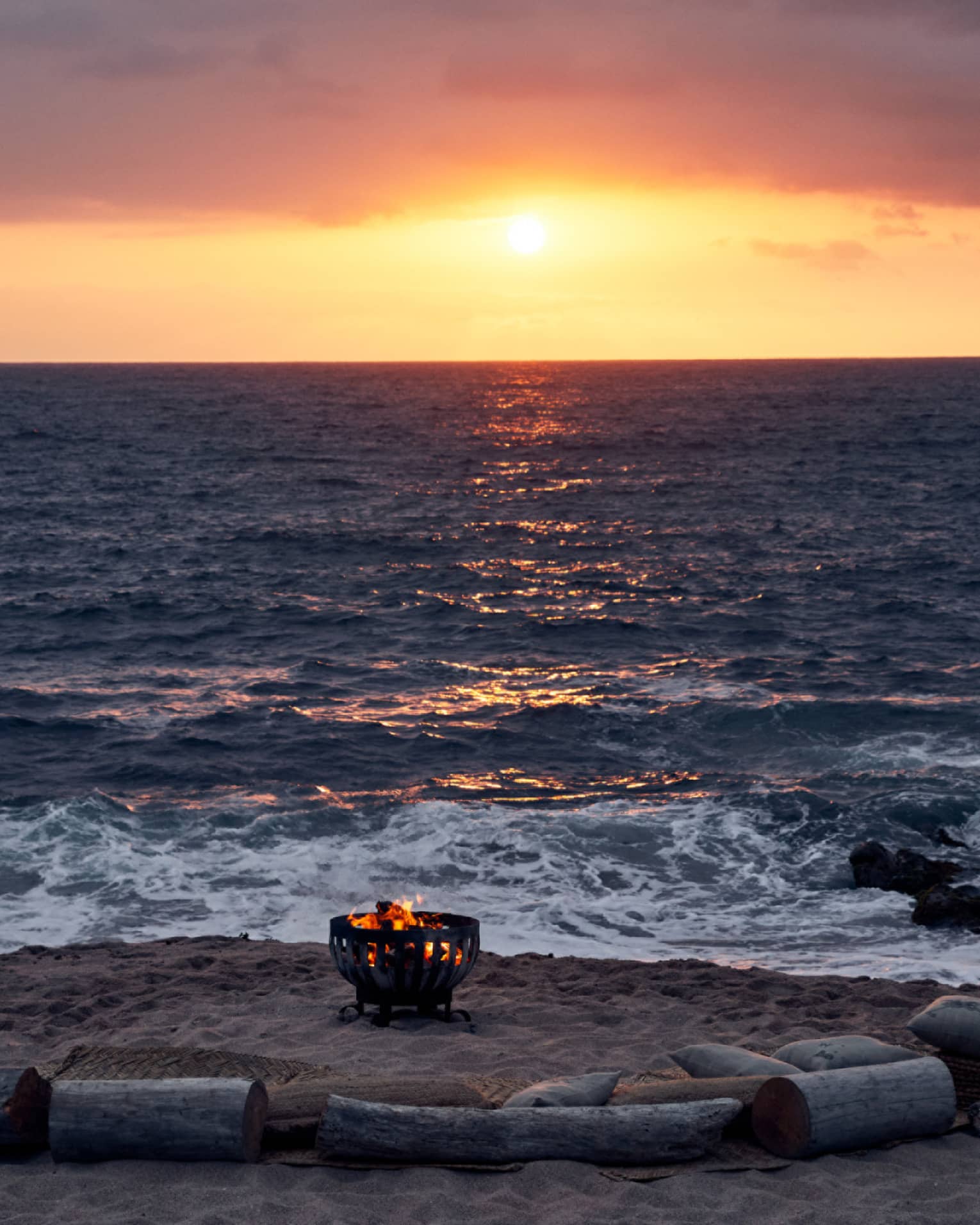 Fire burns in an iron pit on the beach, semi-encircled by driftwood log seats facing the sun setting into the ocean horizon.