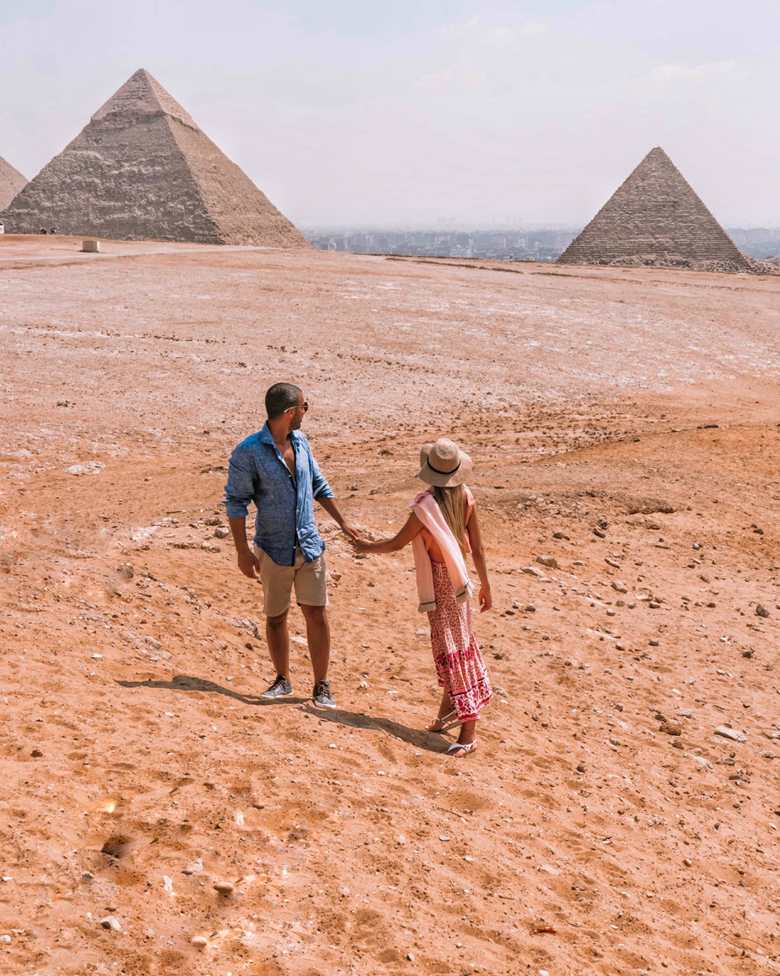 A couple holding hands turn back to look across the arid desert at the giant pyramids in the distance. 