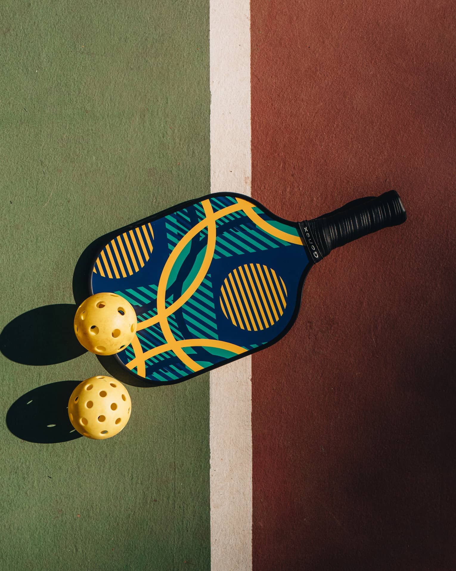 A colourful pickleball racquet and two yellow pickleballs cast shadows upon a green and red court with a white dividing line.