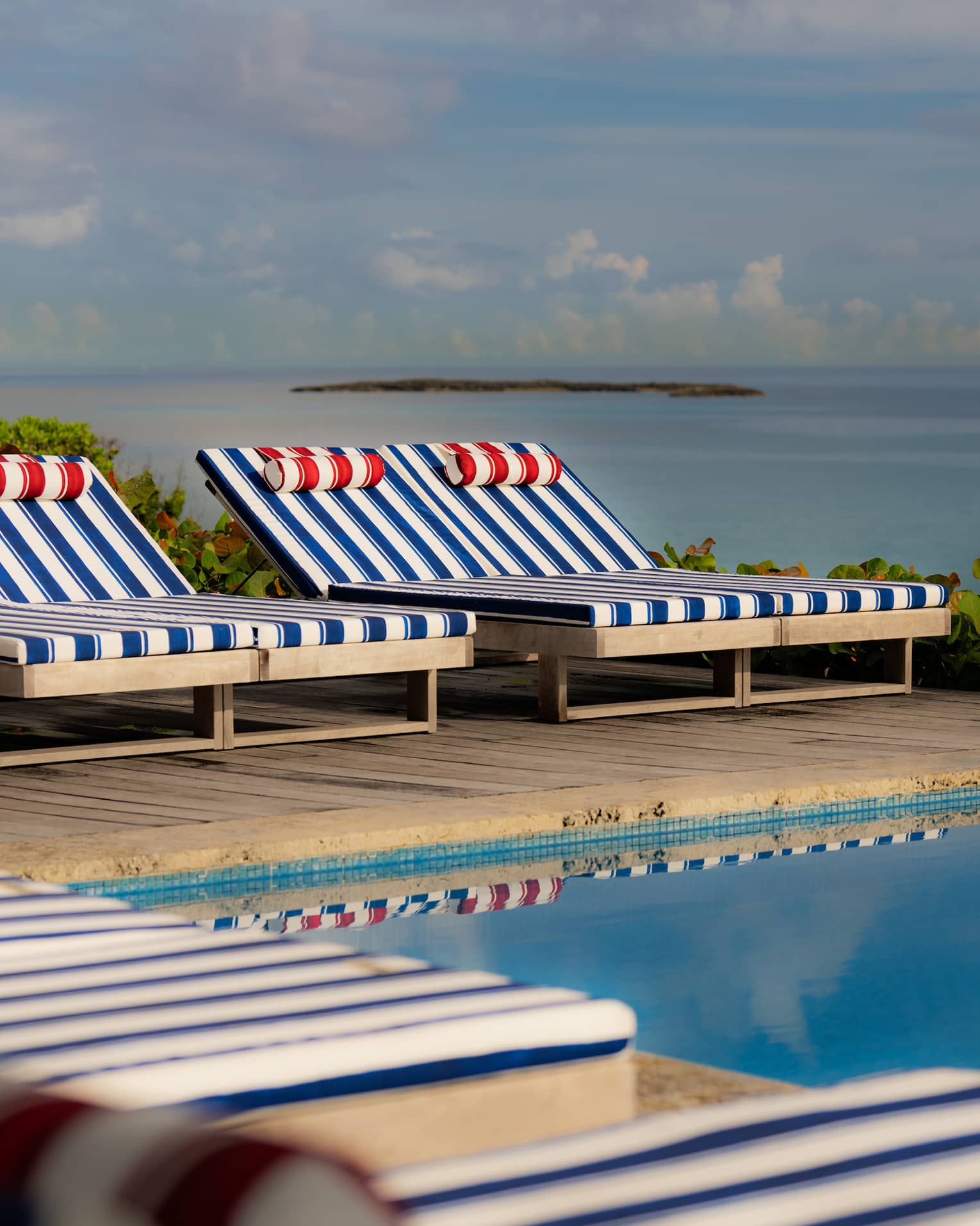 A neat row lounge chairs with blue-and-white striped upholstery and red-and-white striped pillows sit on an elevated pool deck with the ocean in the background