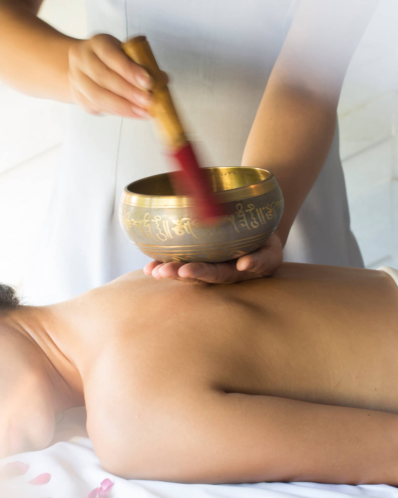 Hands play brass singing bowl over bare back of woman lying on massage table