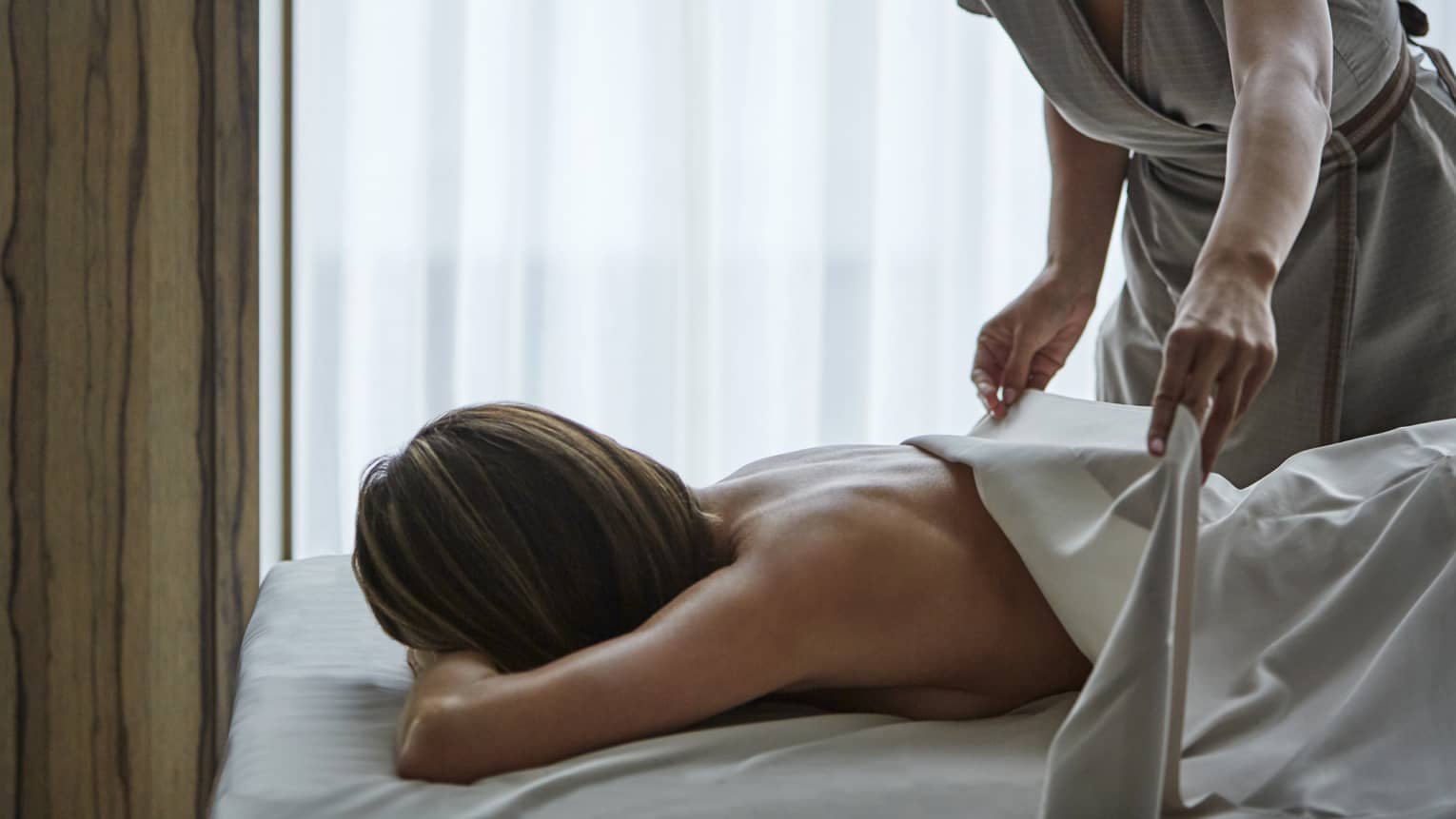 A masseuse covers a woman's back with a sheet during a massage. 
