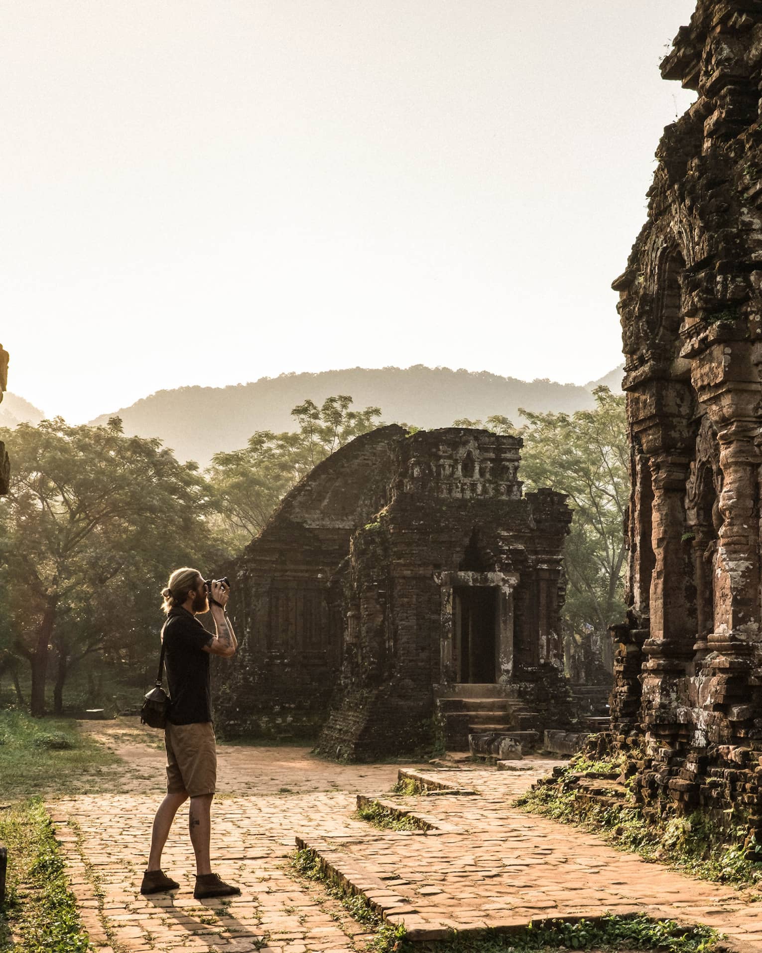 Tourist captures serene beauty of ancient Hindu temple ruins adorned with moss and greenery, sun streaming through the mist.