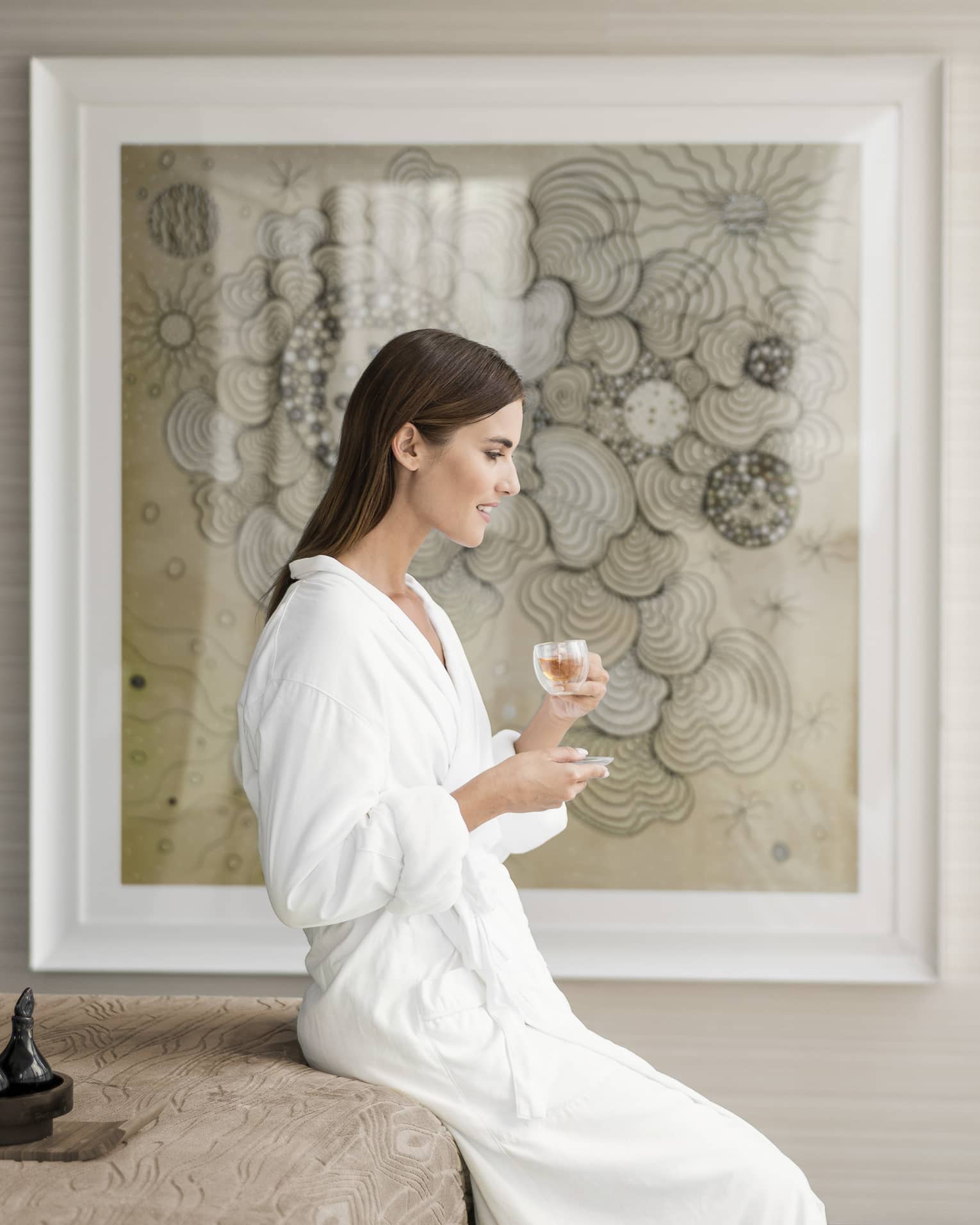 A woman in a white robe sipping from a small glass in front of a framed portrait.