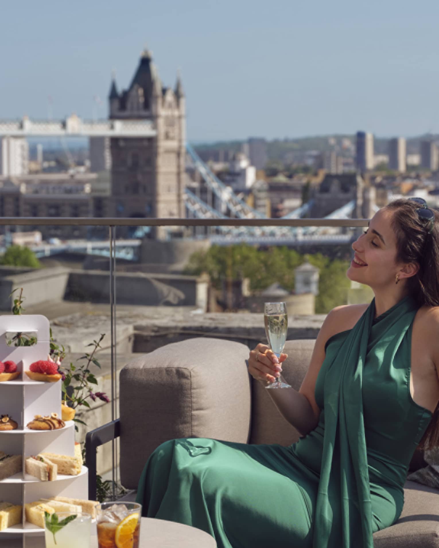 Woman in green dress holding champagne beside tea stand with confections and Tower Bridge in backdrop