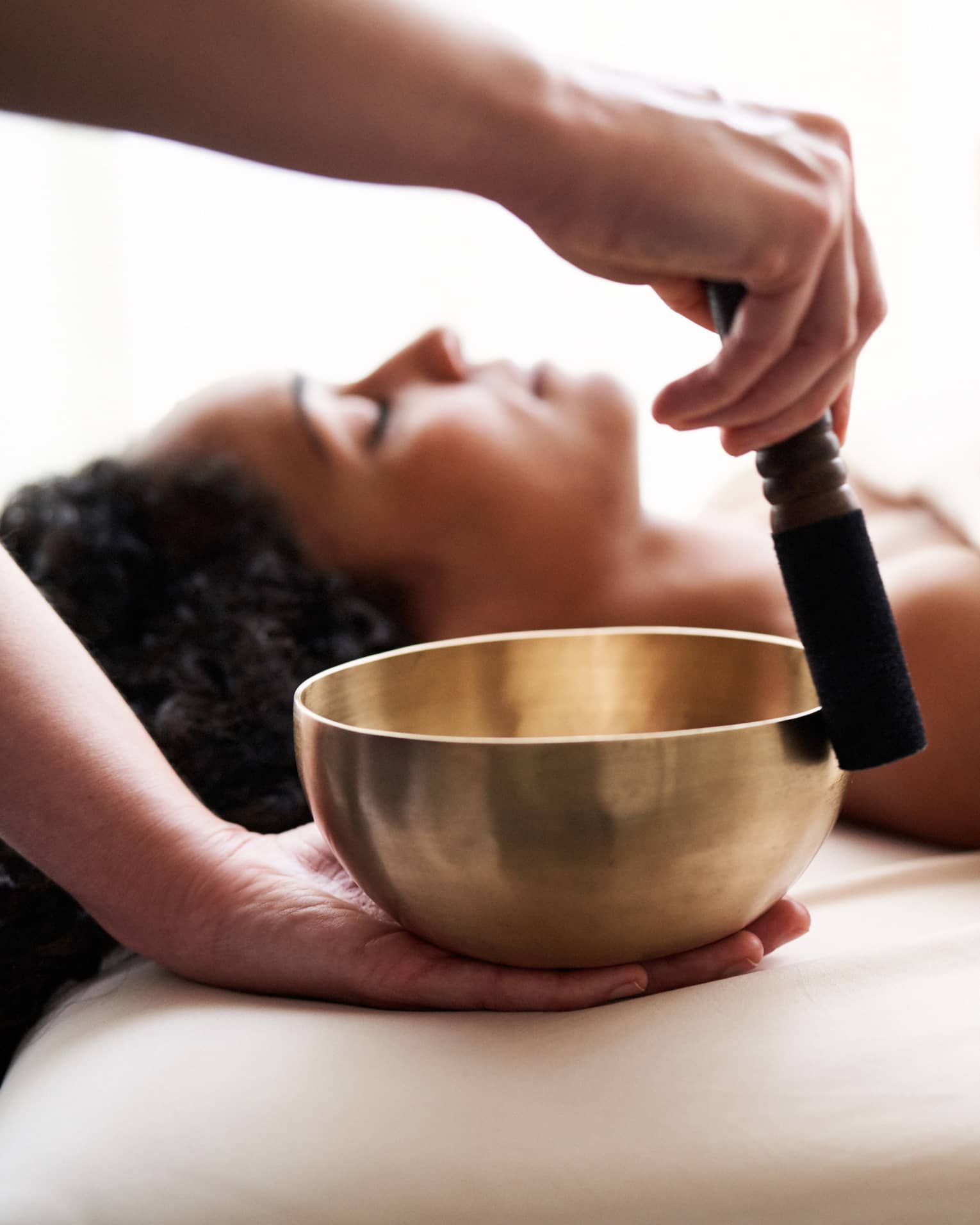 Woman with eyes closed lies on massage table in as vibrational sound therapy is performed with a Tibetan singing bowl