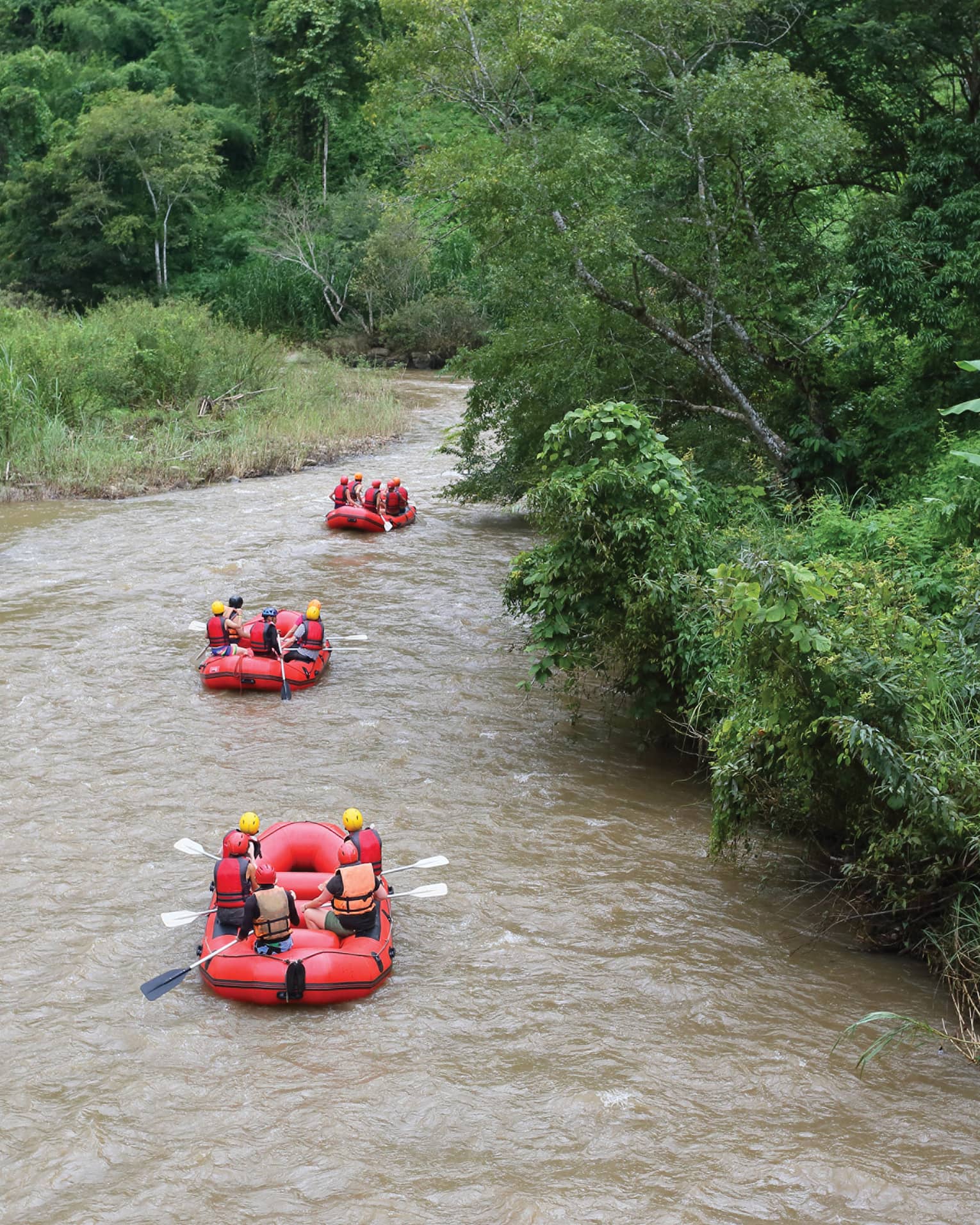 Rear view of three red inflatable rafts, five helmeted riders in each, coasting down a winding river flanked by lush foliage.