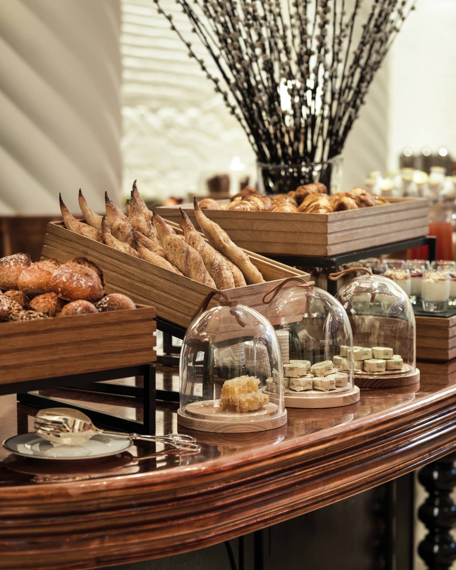 A decadent array of artisan breads and other baked goods in wooden crates and glass-covered dishes on a large wooden table.
