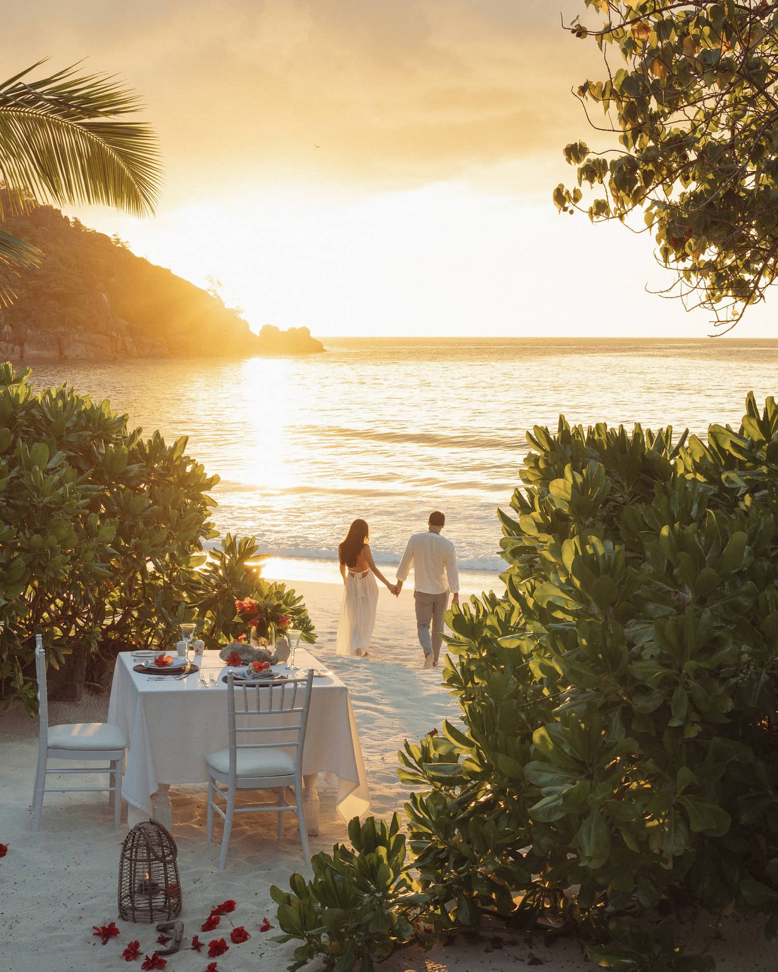 Framed by trees, a romantic dinner table set for two on the beach; a couple holding hands walks in the golden glow of sunset.