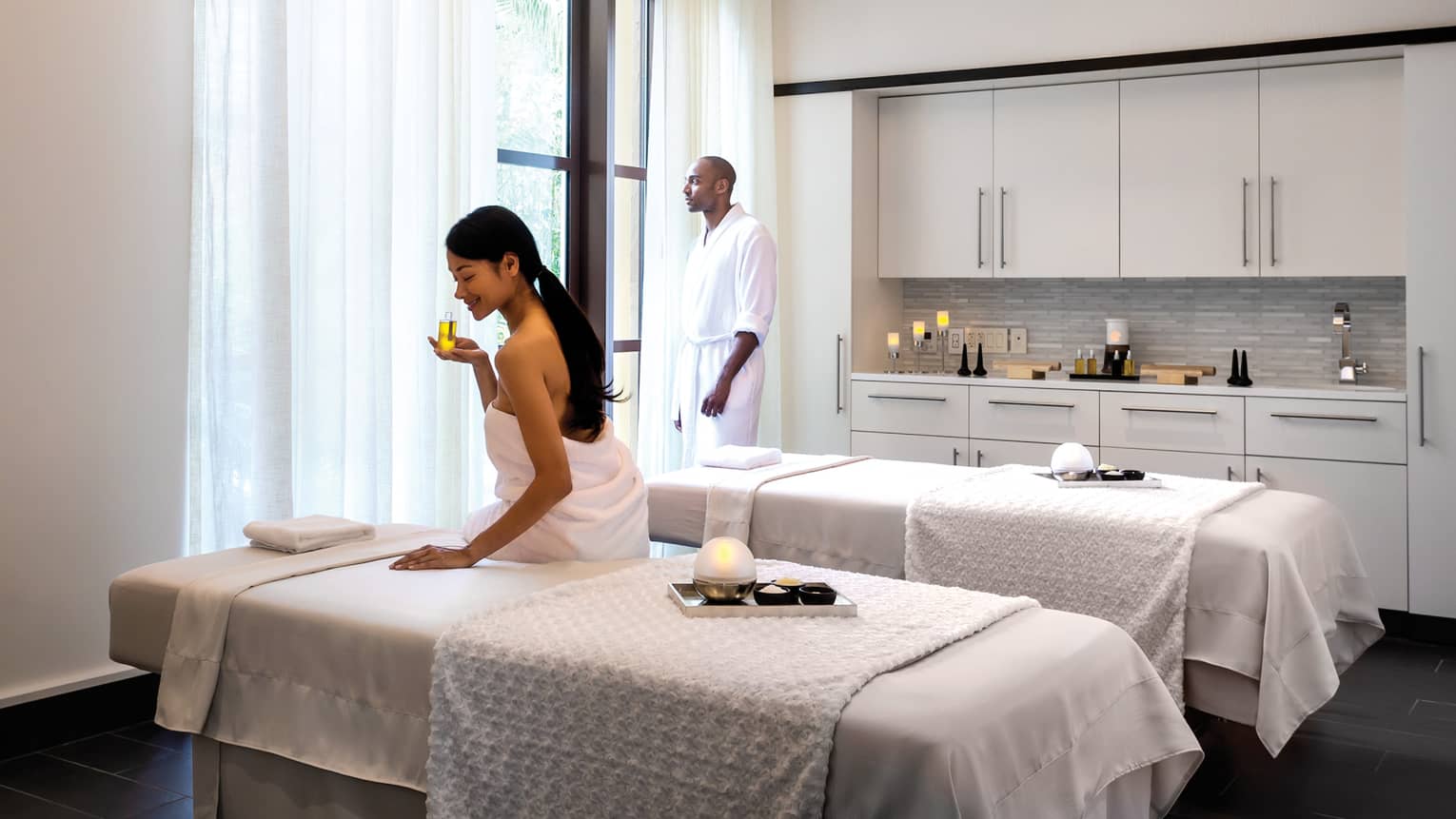 Woman wearing towel sits on edge of Couples Suite massage table, smells oil as man stands at window