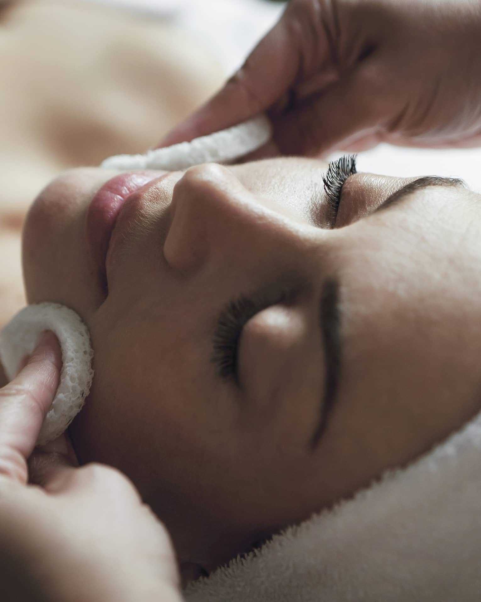 A detail of hands rubbing a sponge on a woman's face as she lies on a massage table with her eyes closed and hair wrapped up in a white towel