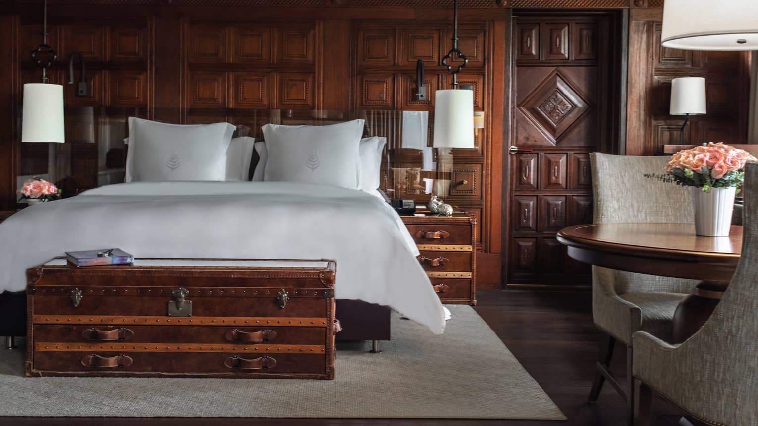 Bedroom with dark wood walls, white king bed, trunk, table with two chairs