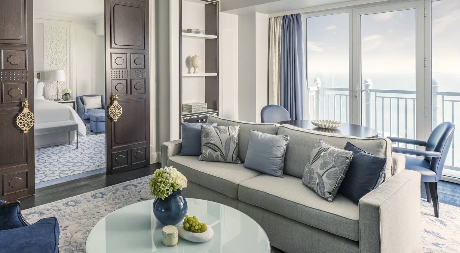 Sea-view living room, with grey sofa, blue pillows and accent chairs, chandelier, dark wooden doors open to bedroom