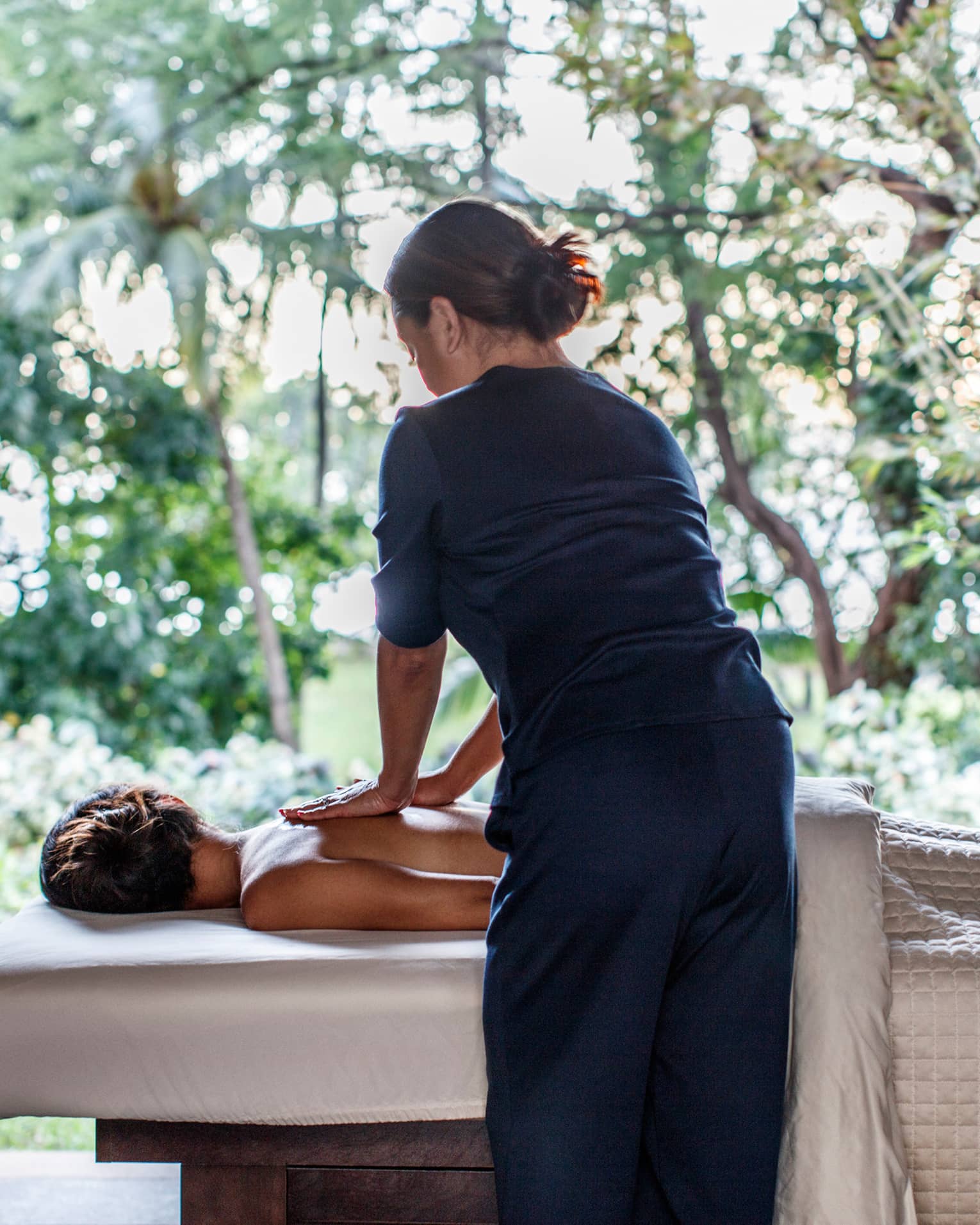 Spa staff massages woman's bare shoulders as she lies under sheet on massage table