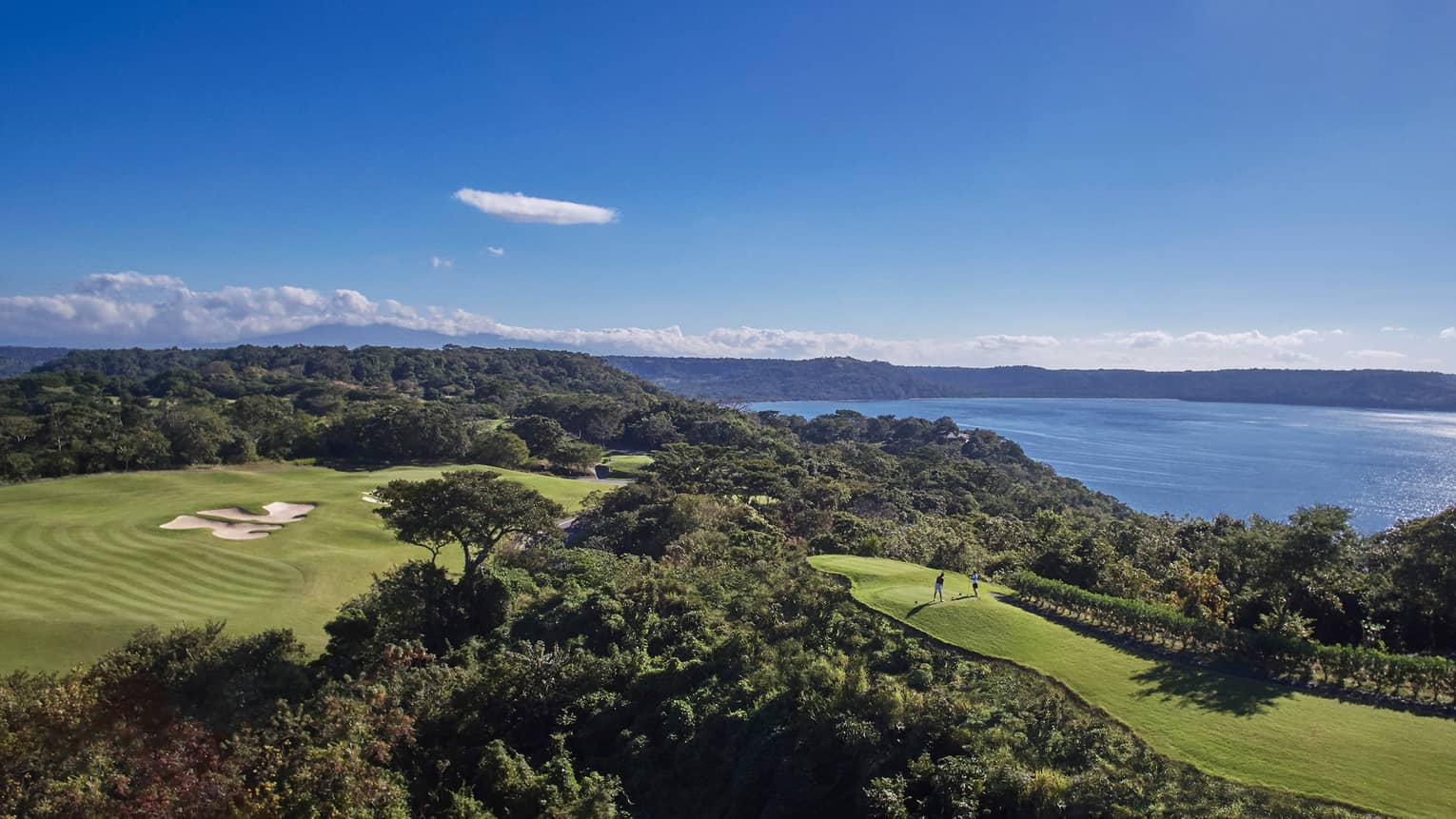 Looking down at large golf course green, trees on top of mountain, two players on hole, ocean view
